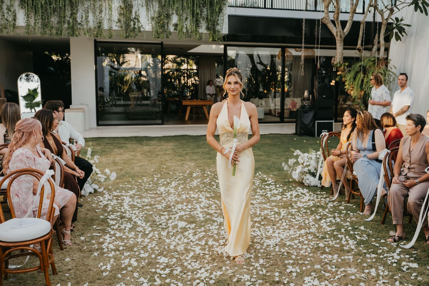 Bridesmaid walks down petal-lined aisle in gold dress carrying bouquet at elegant outdoor wedding ceremony.