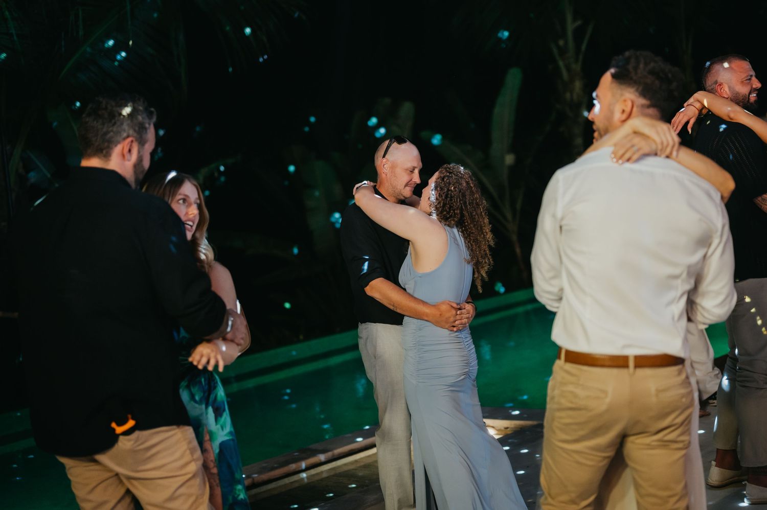Wedding guests dancing and embracing near a glowing pool during an outdoor evening tropical reception.