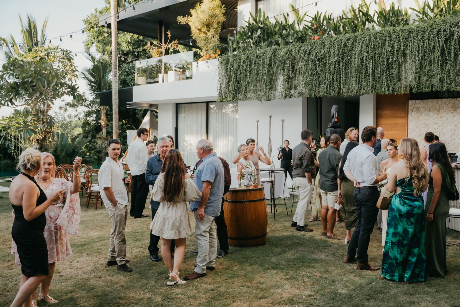 Wedding guests mingling outdoors on lawn in front of modern venue with hanging greenery.