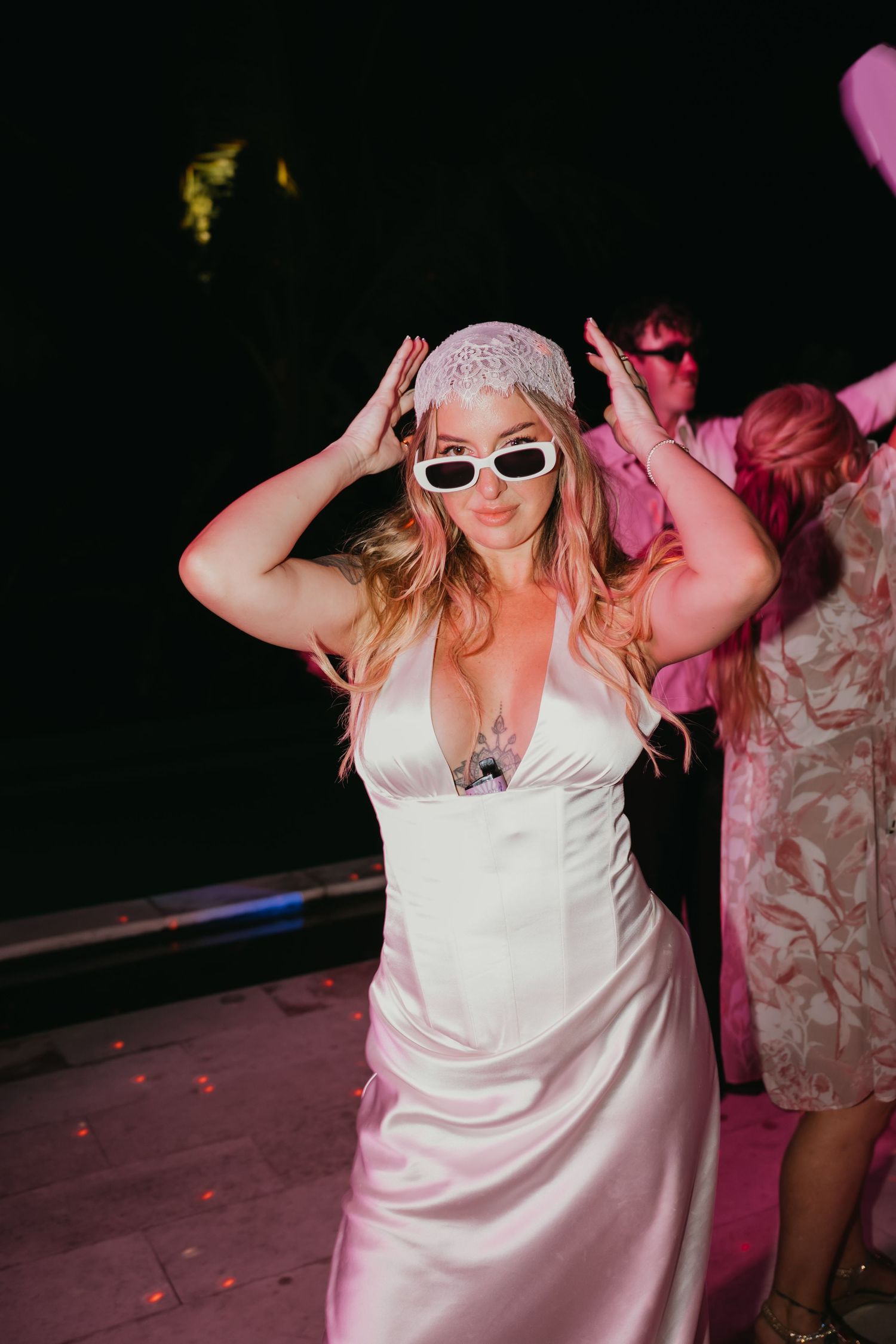 Bride in satin gown wearing white sunglasses and lace headpiece dancing at night reception.