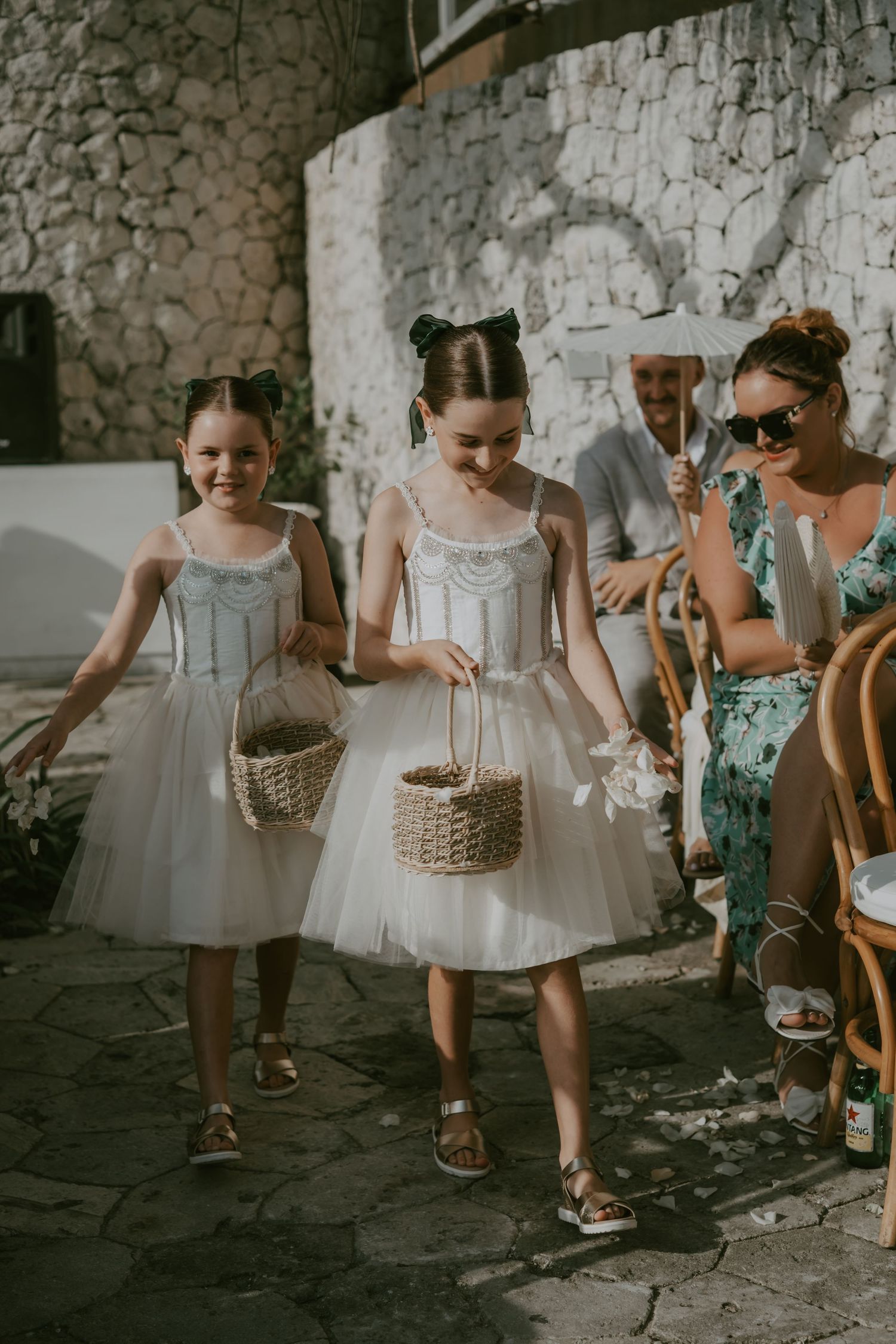 Young attendants in white dresses carry woven baskets along a stone pathway at an outdoor wedding.