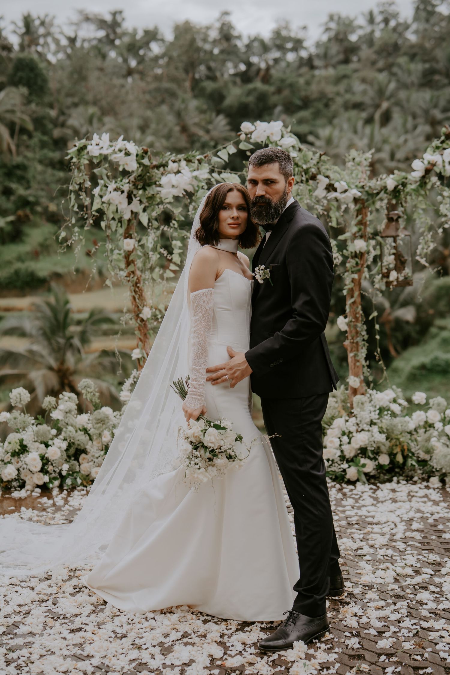 A romantic wedding couple poses together in a garden setting surrounded by delicate white floral arrangements.