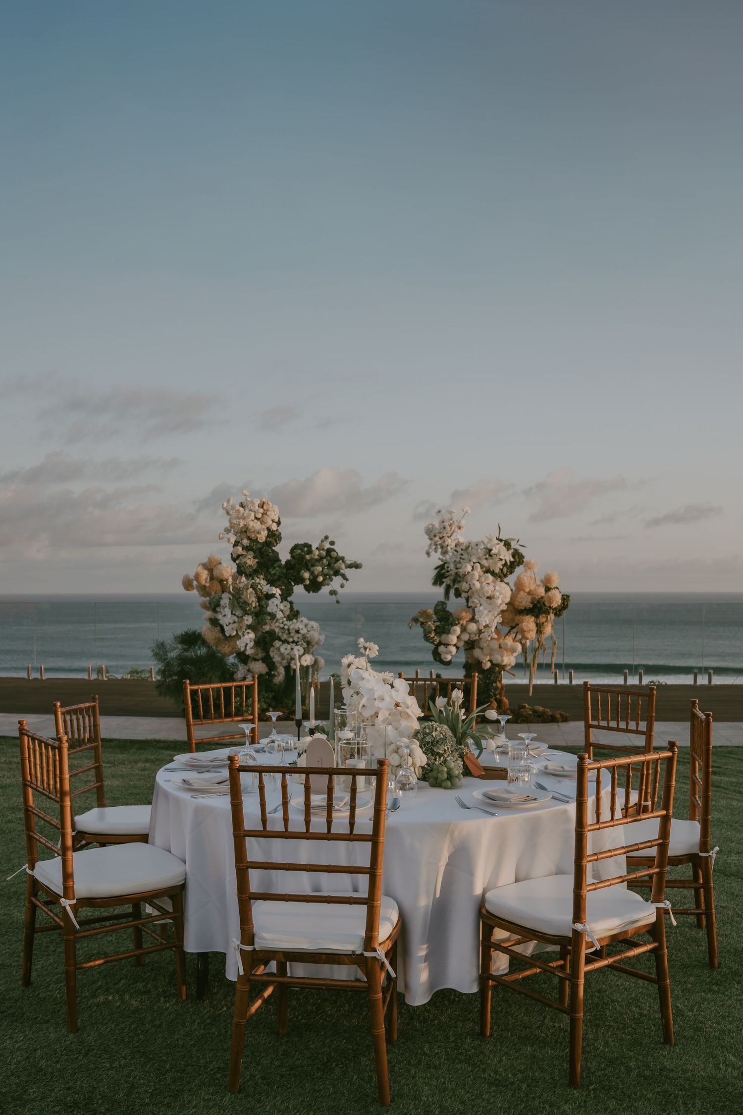 Elegant outdoor wedding reception setup with wooden chairs and white linens overlooking an ocean view at sunset.