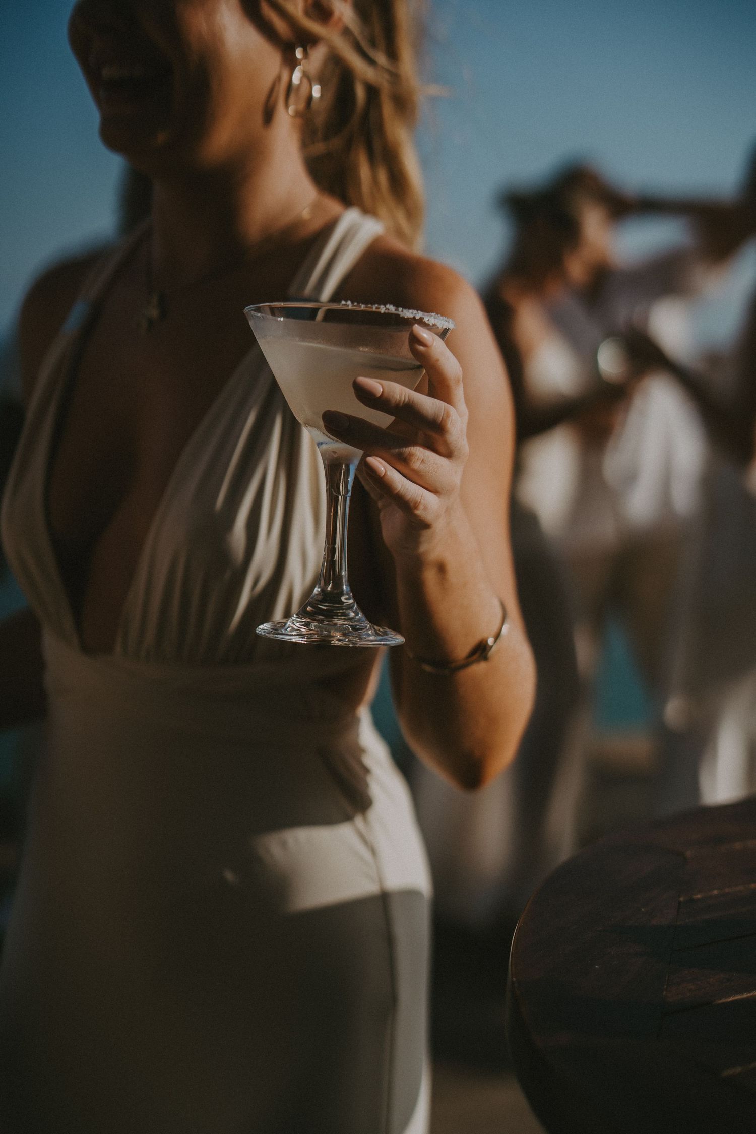 Person in elegant dress holds martini glass with sugar rim at sunset while others gather in background at outdoor event.
