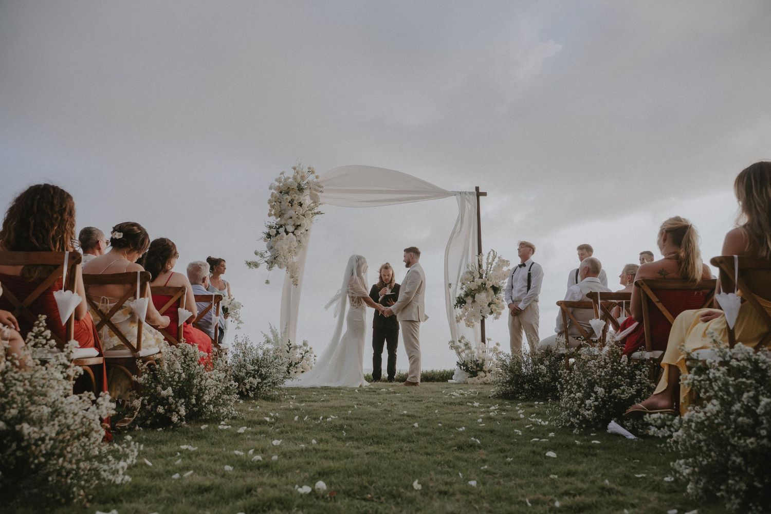 Wedding ceremony guests gather under cloudy skies at an outdoor hilltop venue with white tent and floral arrangements.