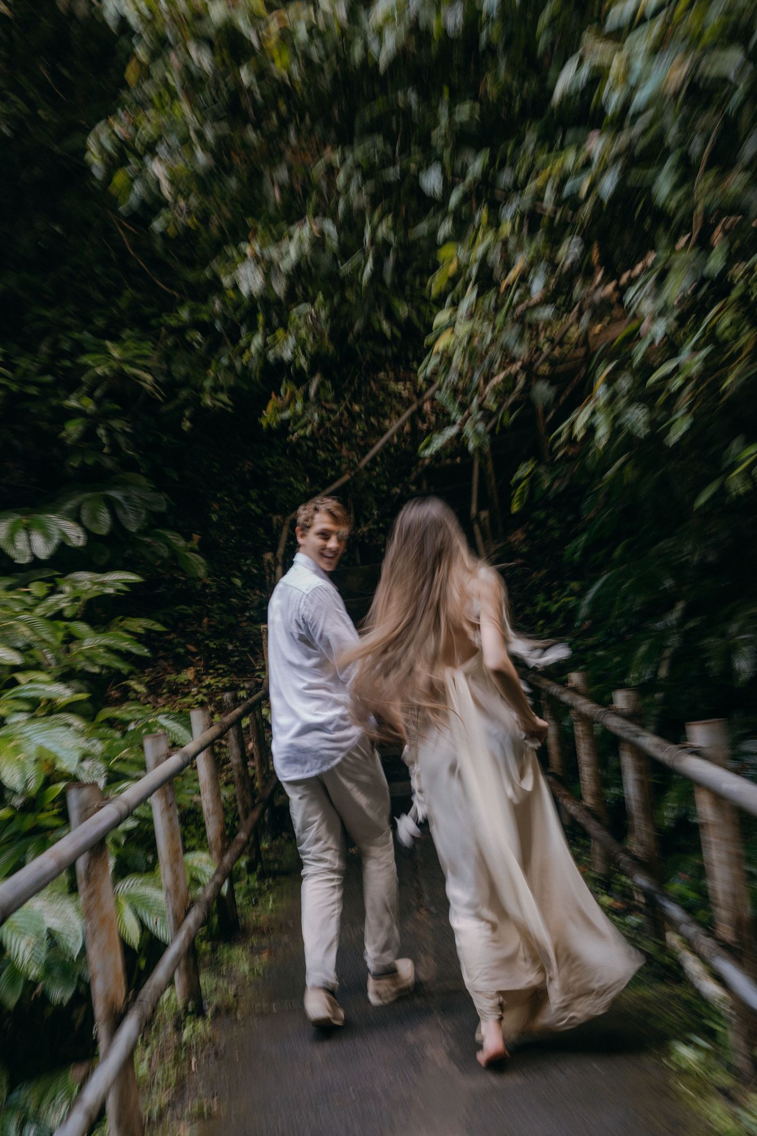 A couple in white outfits walks along a wooden pathway surrounded by lush greenery and tropical plants.