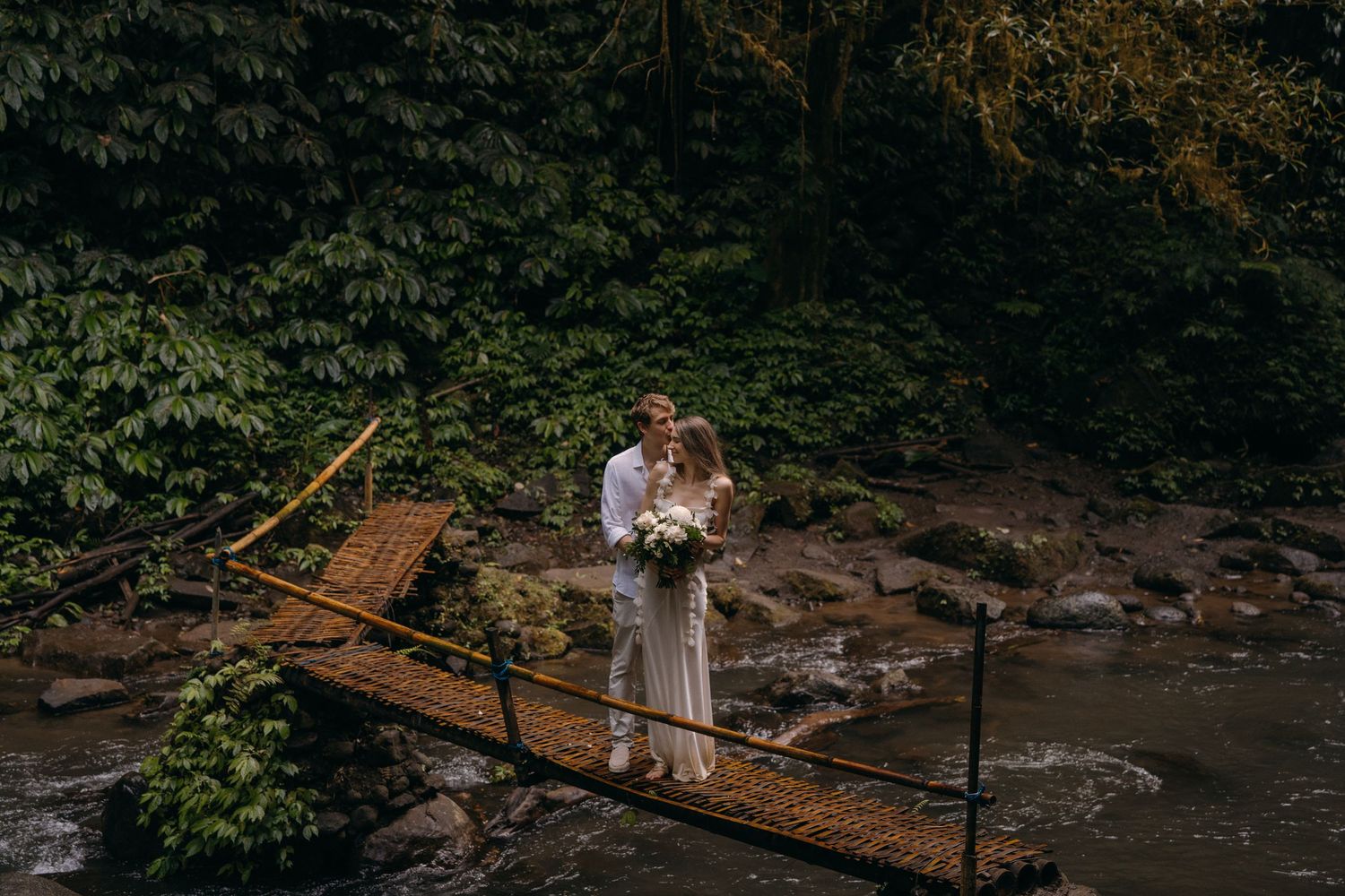 Couple standing on wooden bridge in tropical forest setting with lush greenery and flowing stream below.
