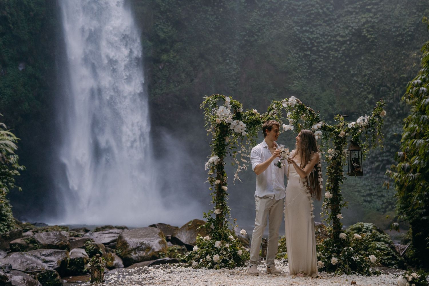 A couple shares a romantic moment under a floral arch with a majestic waterfall cascading in the background.