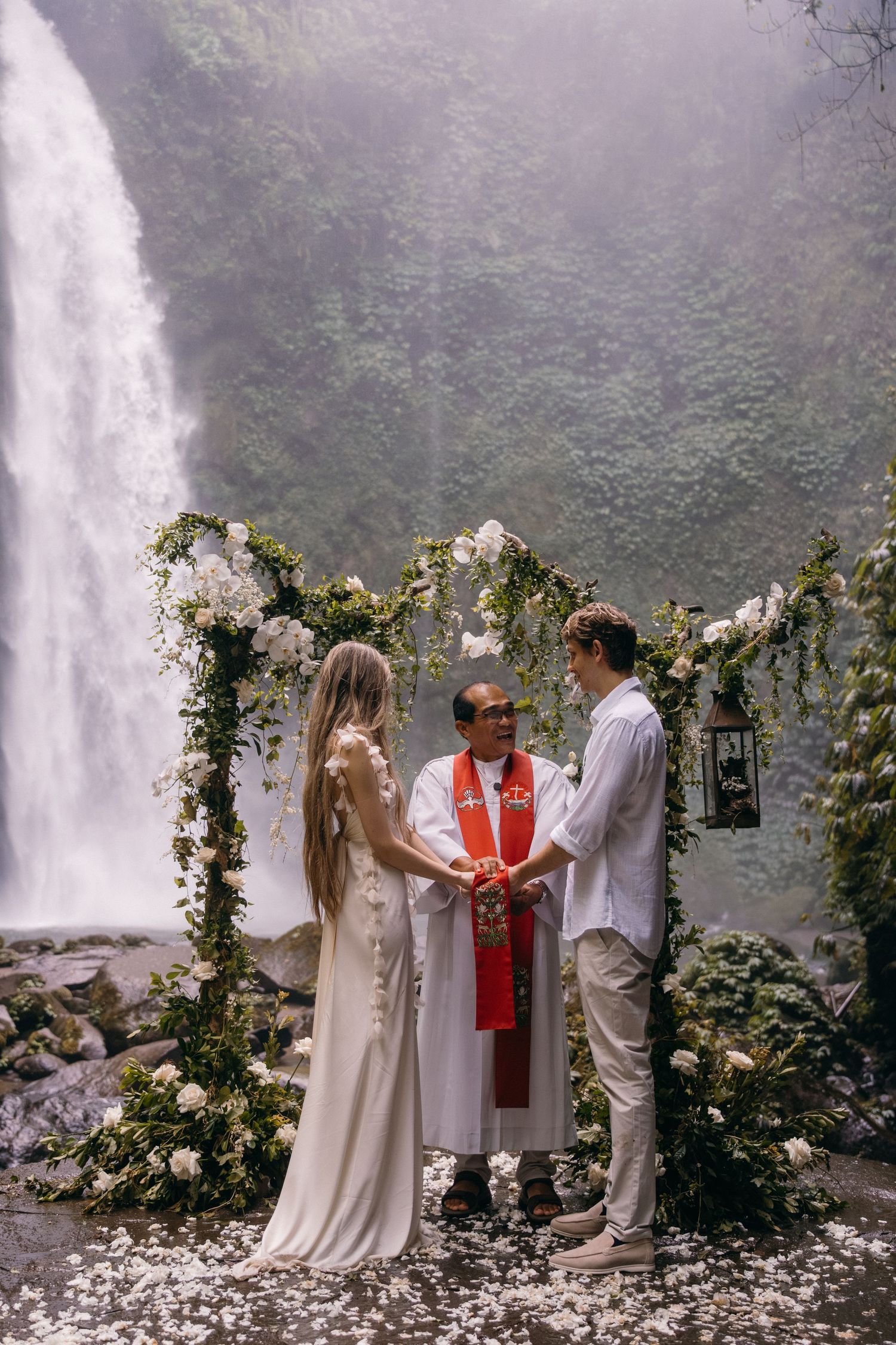 A wedding ceremony takes place in front of a misty waterfall with floral decorations and white-robed figures.