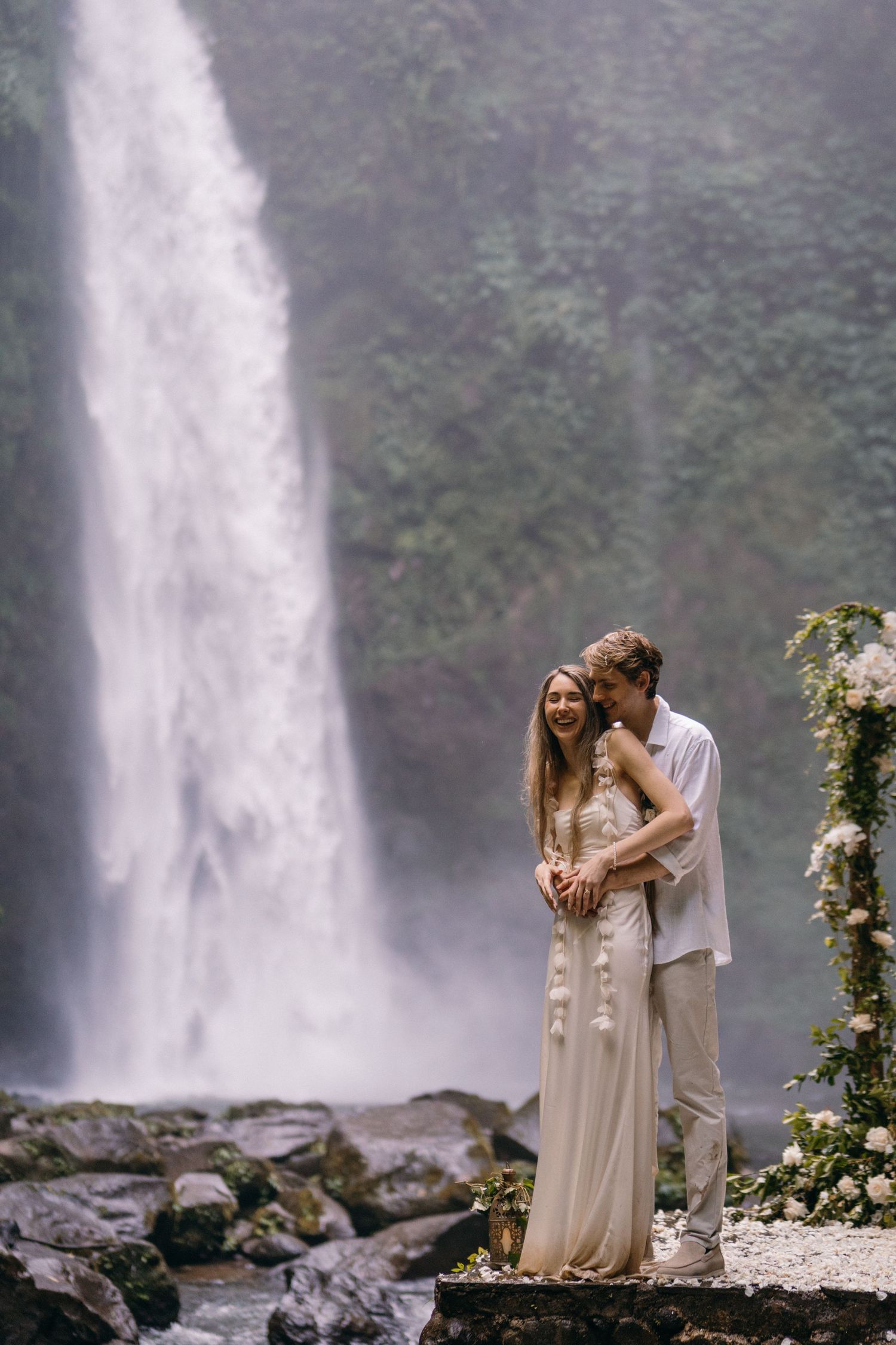 A romantic couple stands by a majestic waterfall surrounded by misty cliffs and floral decorations in a wedding setting.