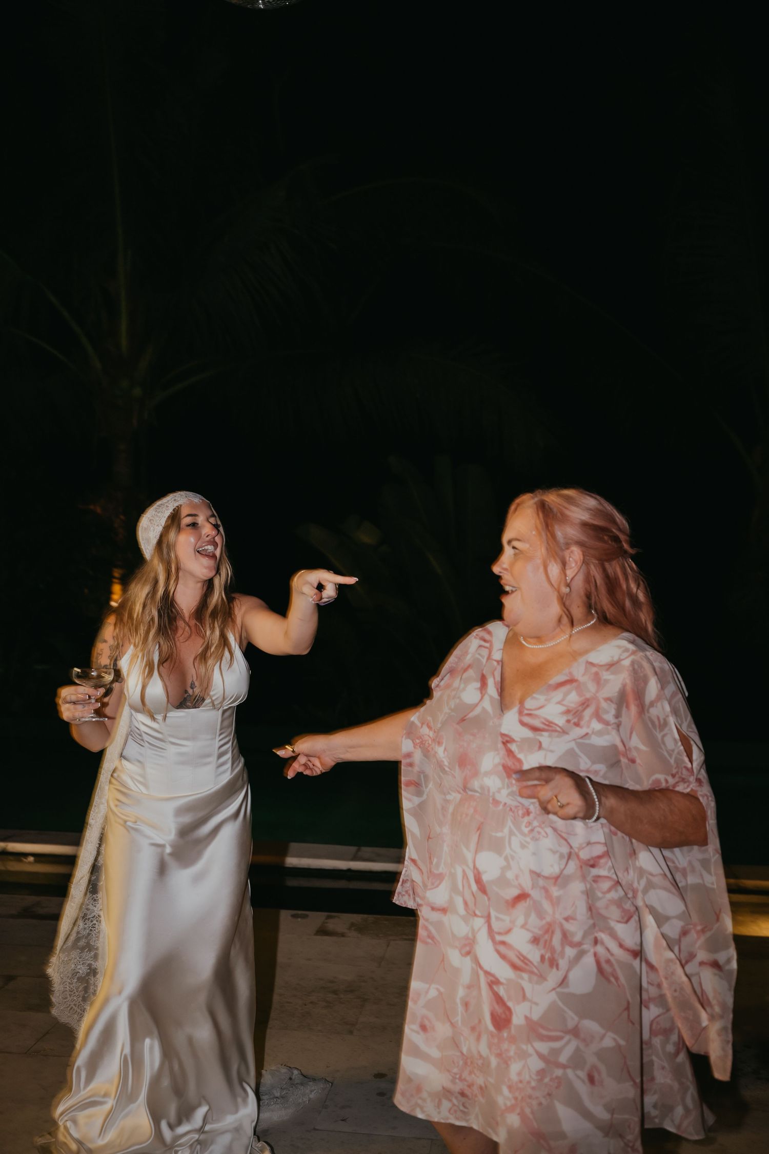 Bride in white satin gown and headband dancing and laughing with a guest in a pink floral dress at night.