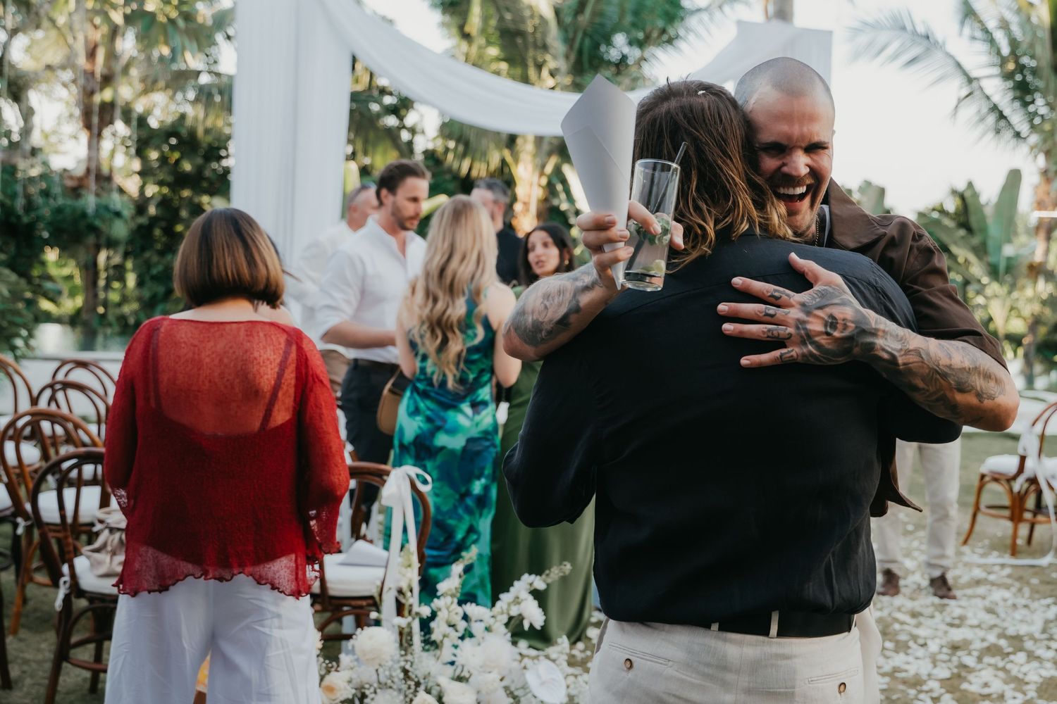 Tattooed groom sharing a joyful hug with a guest while holding champagne glasses after the ceremony.