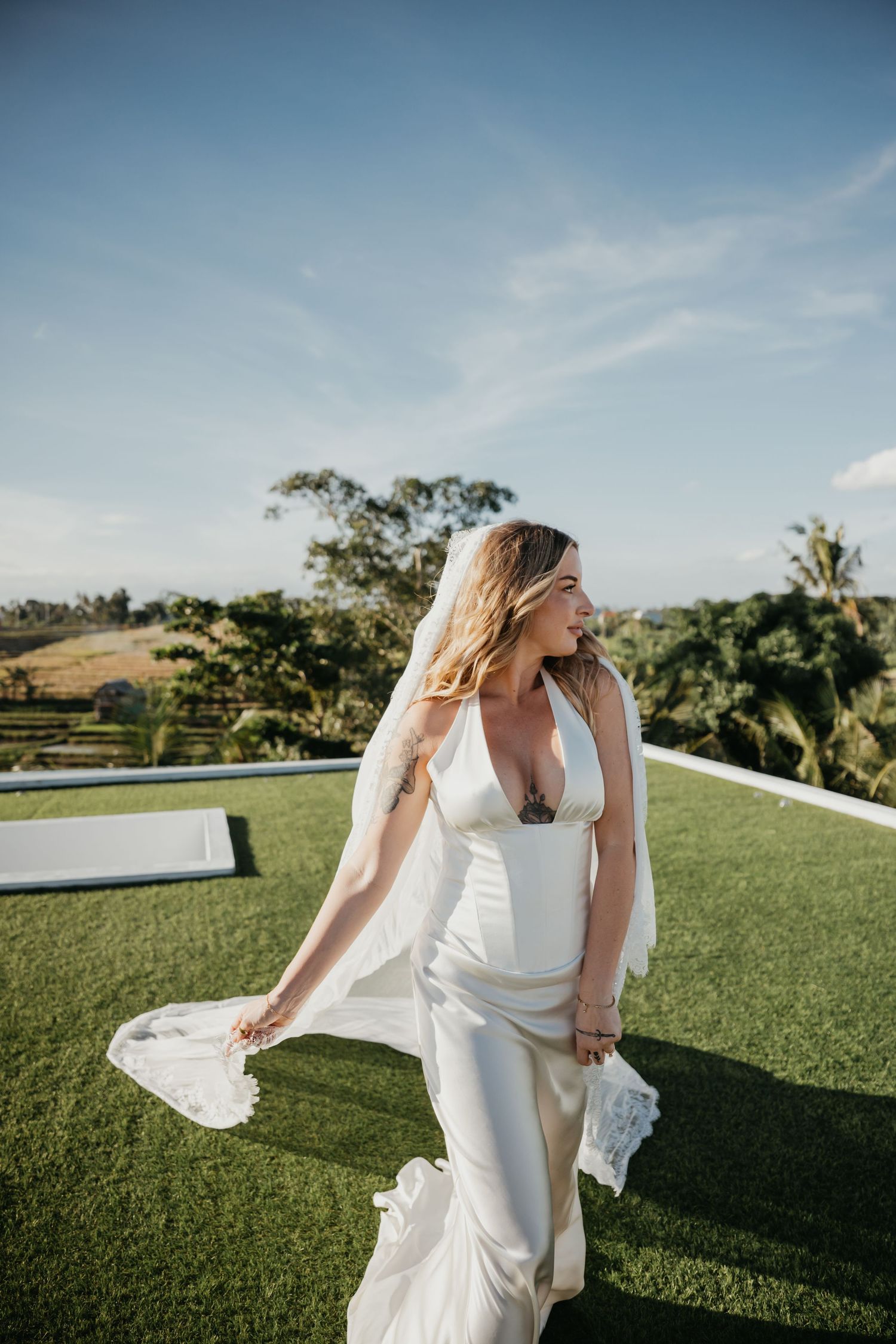 Blonde bride in elegant white satin gown with veil flowing in breeze standing on green lawn with tropical scenery.