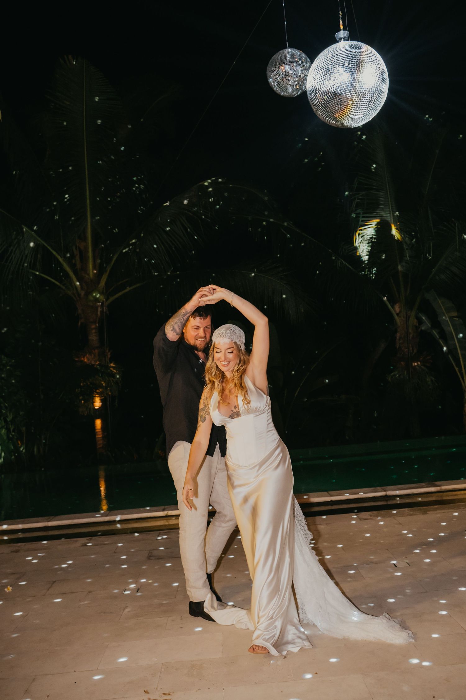 Bride and groom share first dance under disco balls at night with palm trees and confetti in background.