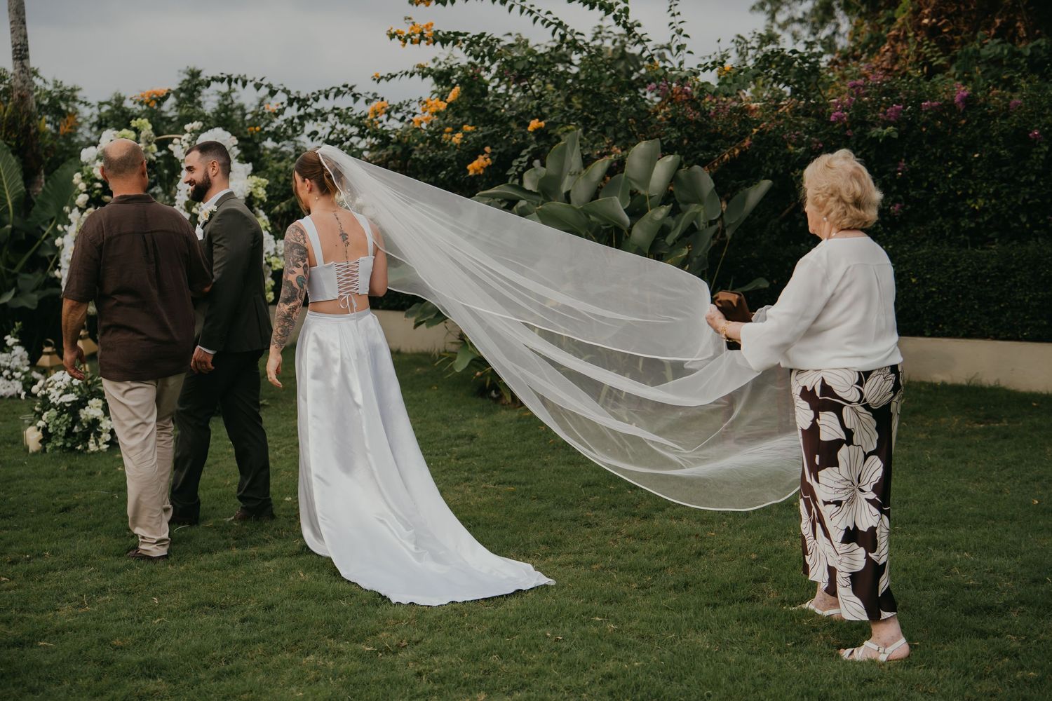 Bride in a white gown with flowing veil walking toward the altar with family during outdoor ceremony.