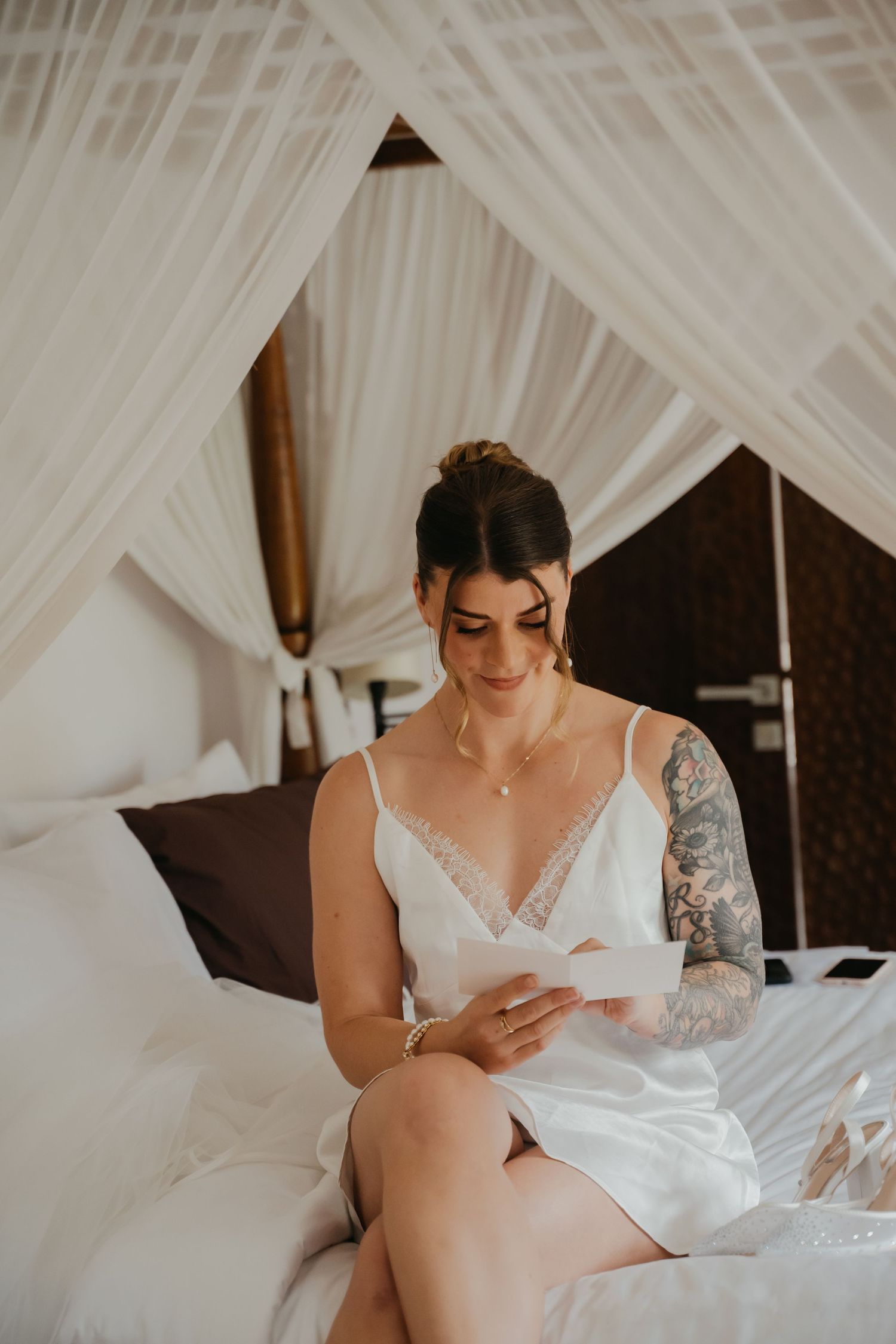 Tattooed bride in white lace slip reads a letter while seated on elegant canopy bed before wedding.