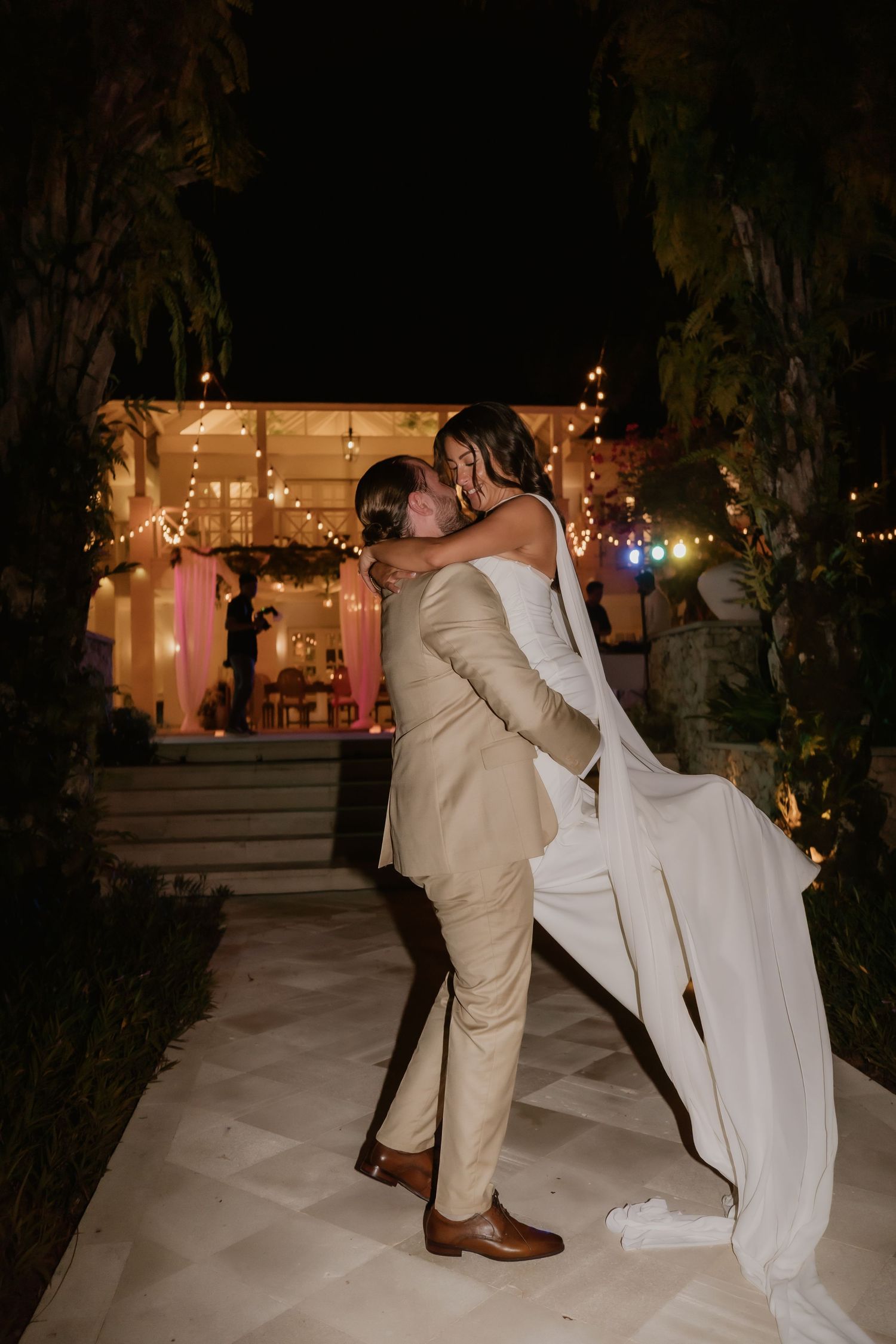 Groom lifting bride for a kiss under string lights outside wedding venue.