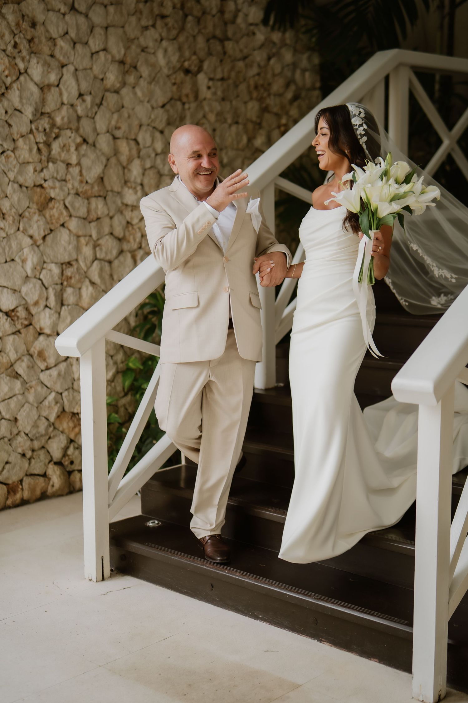 Bride and father descending stone staircase laughing together before outdoor tropical wedding ceremony.