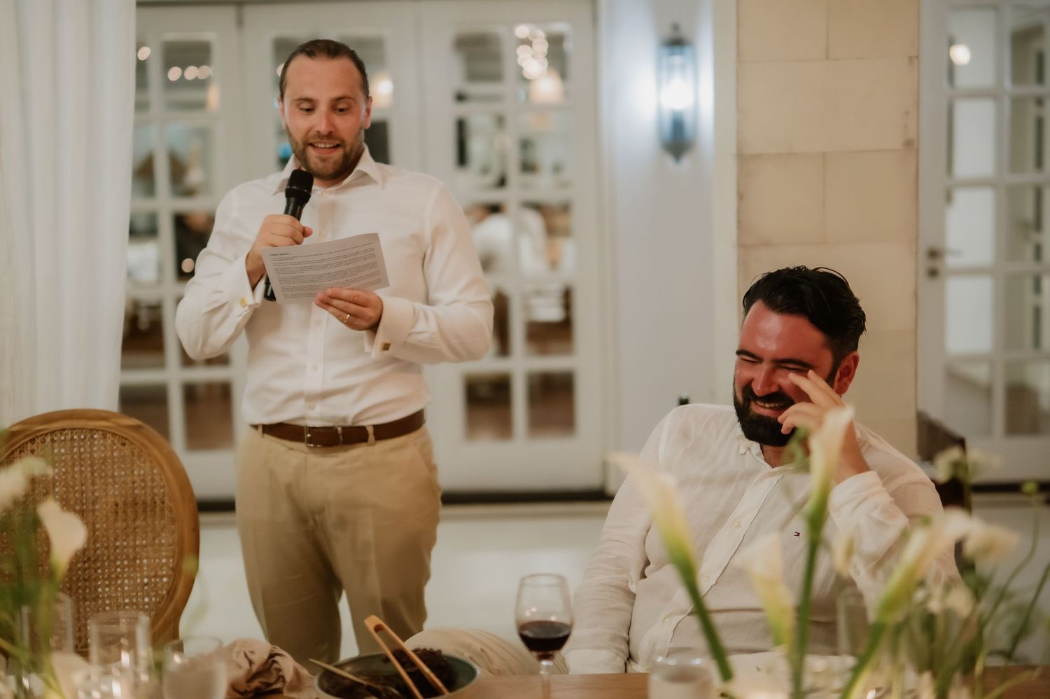 Best man delivers heartfelt speech while groom laughs emotionally at elegant wedding reception dinner.