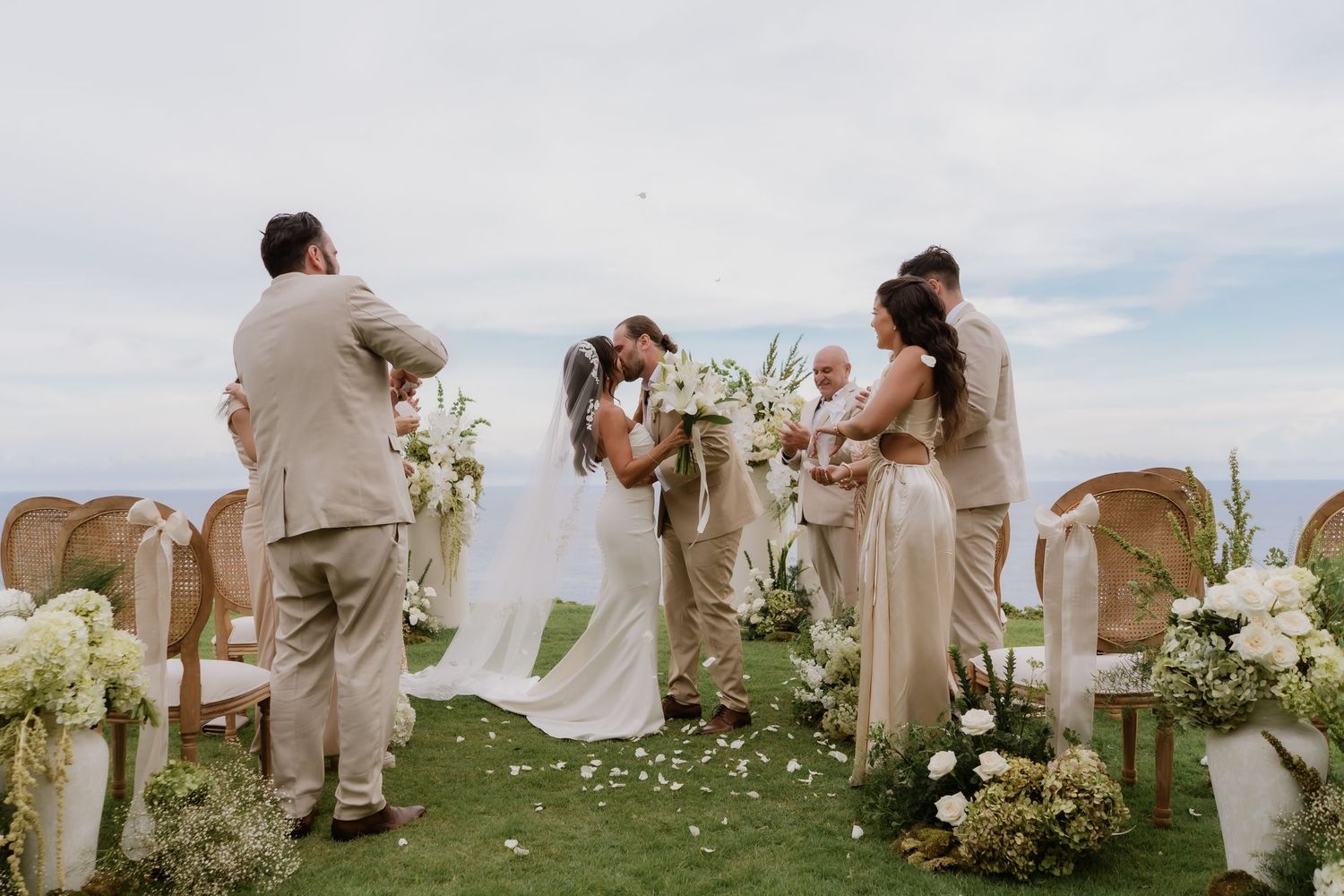 Couple shares first kiss at outdoor wedding ceremony with white floral arrangements and ocean view.