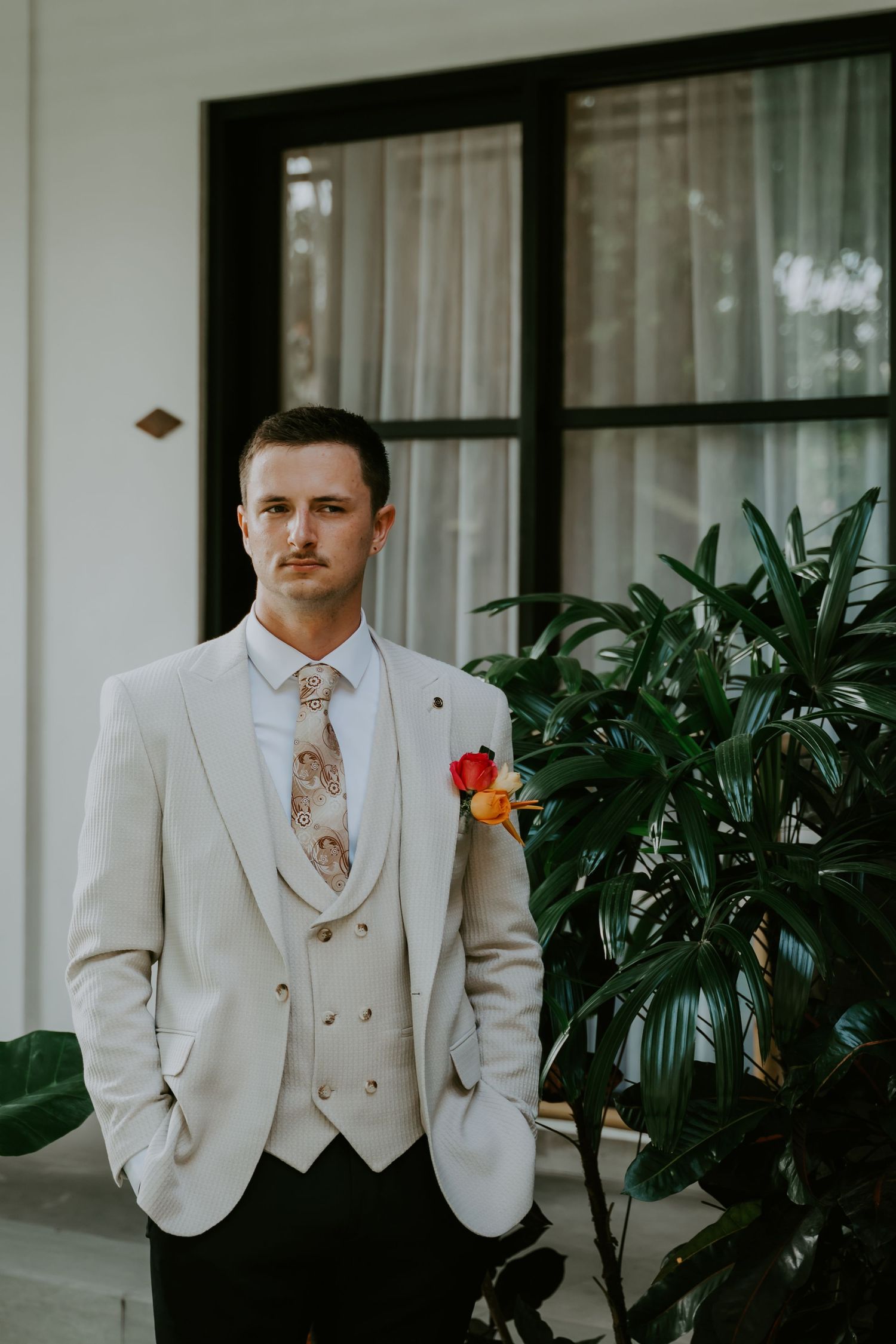 Groom in cream three-piece suit with floral boutonniere poses before large window with tropical plants.