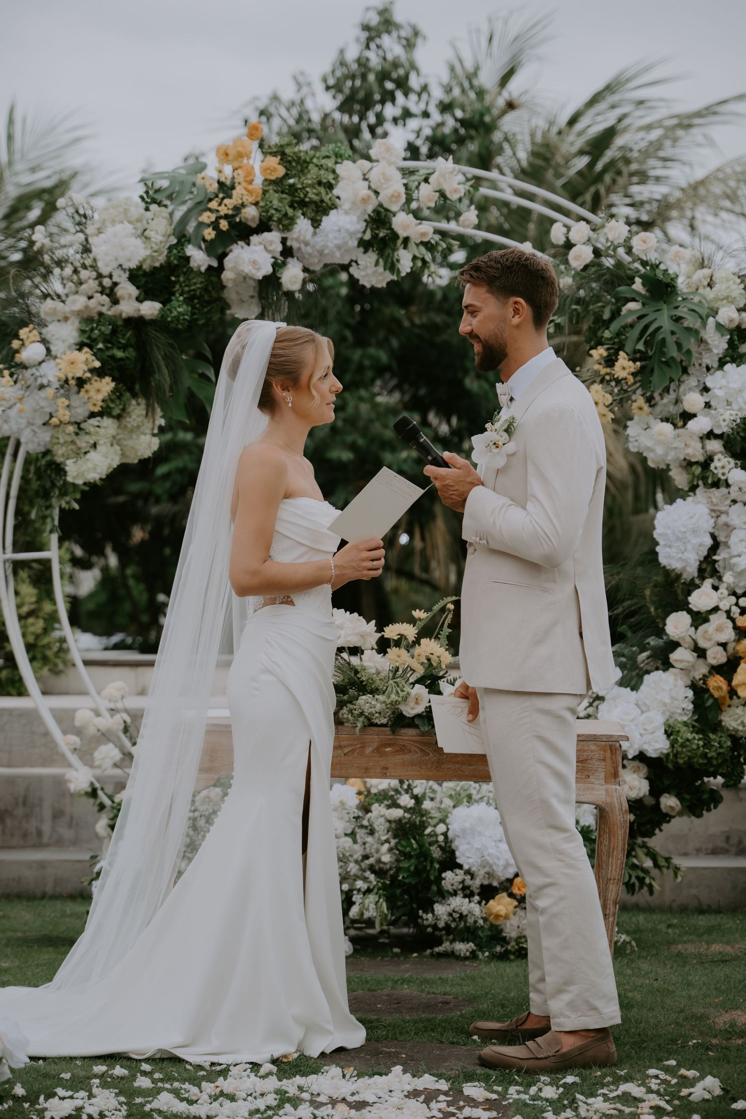 Bride and groom exchange vows under floral arch with white and yellow blooms at outdoor ceremony.