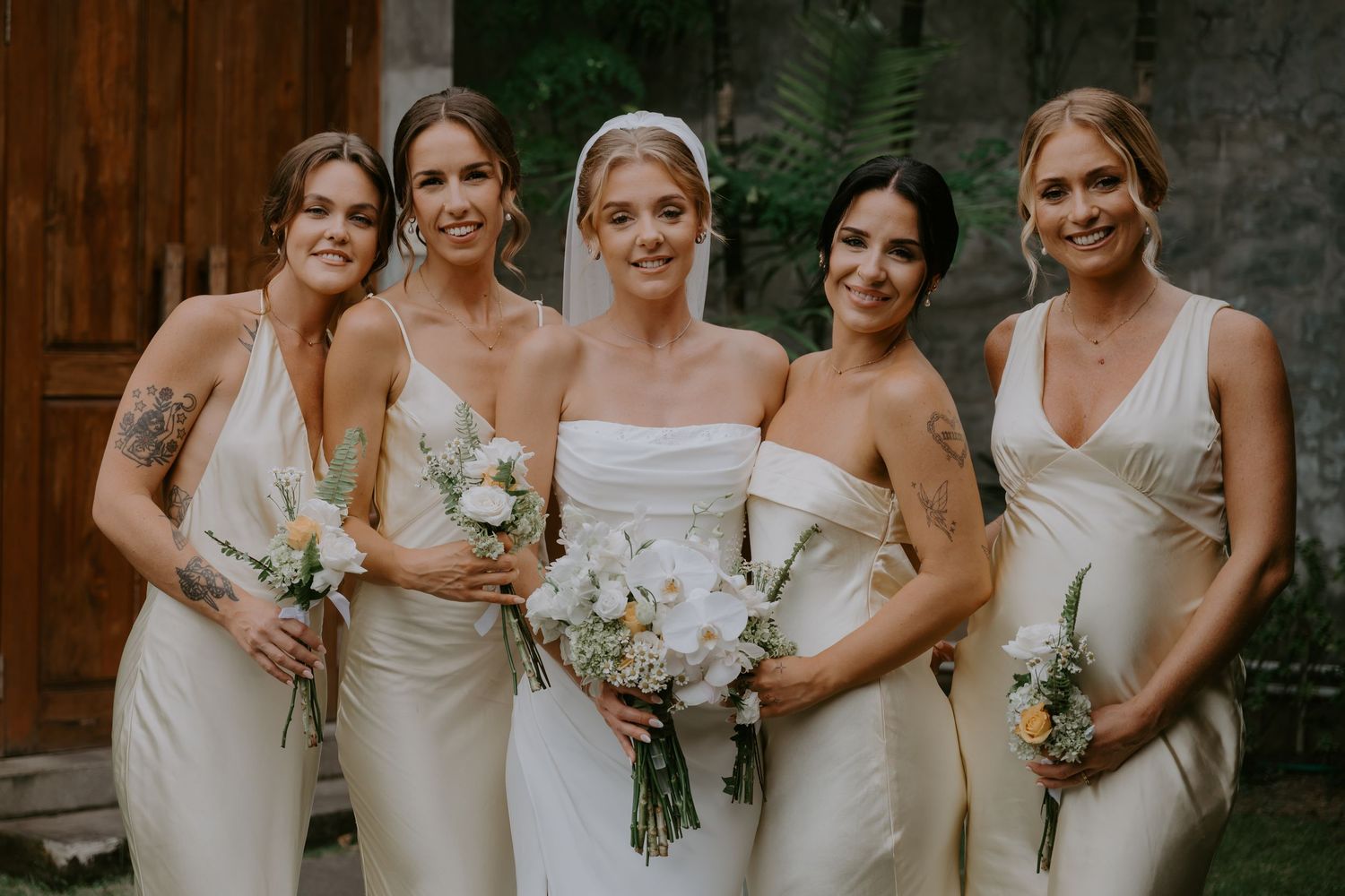 Bride in strapless white gown with three bridesmaids in cream dresses holding floral bouquets.