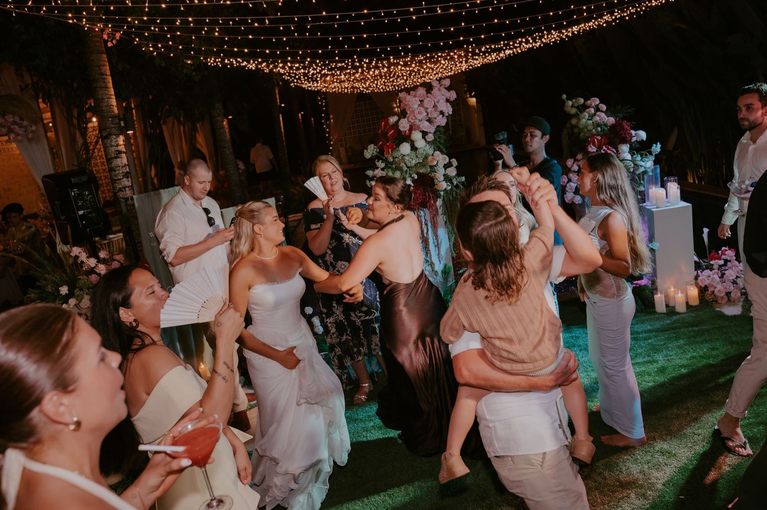 Bride dancing joyfully with wedding guests under fairy lights on outdoor reception dance floor.
