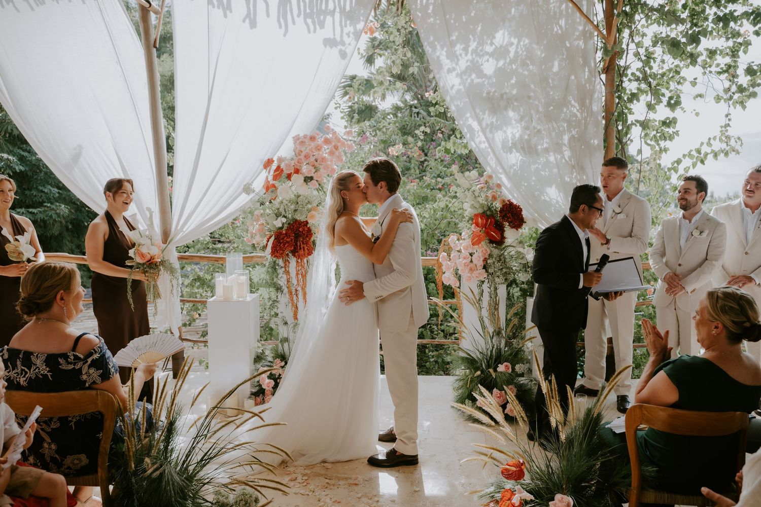 Bride and groom share first kiss at tropical outdoor wedding ceremony with floral arch and draped white fabric.