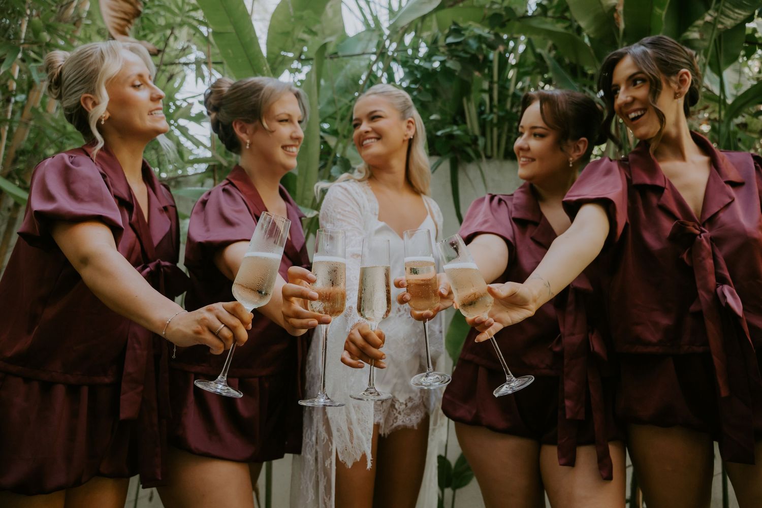 Bridesmaids in burgundy satin sets toast champagne flutes with the bride in a white lace outfit outdoors.