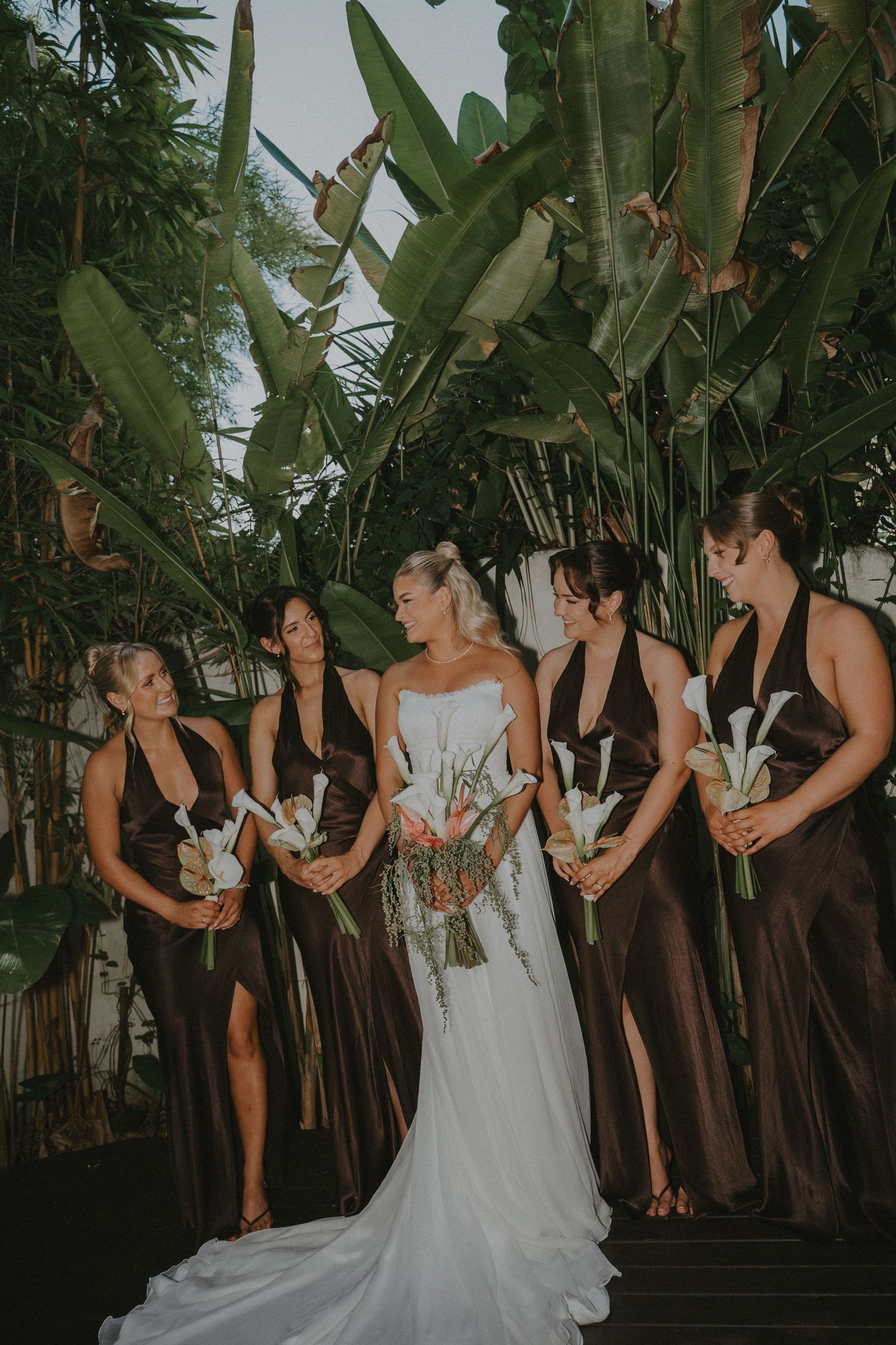 Bride in white gown with four bridesmaids in dark brown dresses holding white calla lily bouquets among tropical foliage.