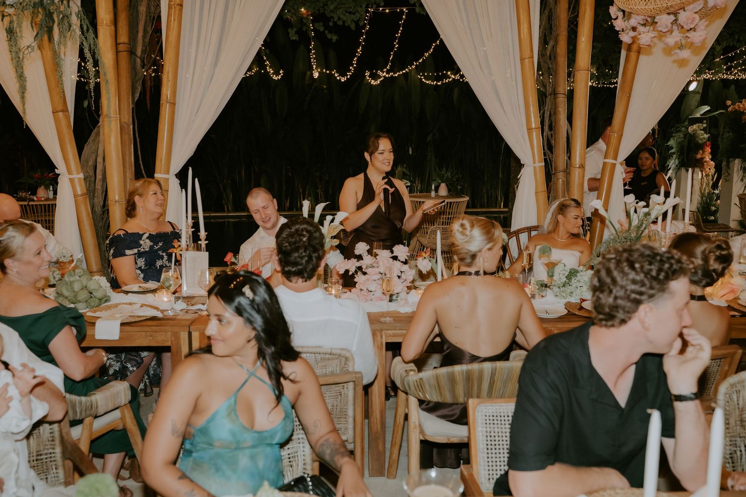Wedding guests seated at decorated tables listen to a speech during an outdoor evening reception.