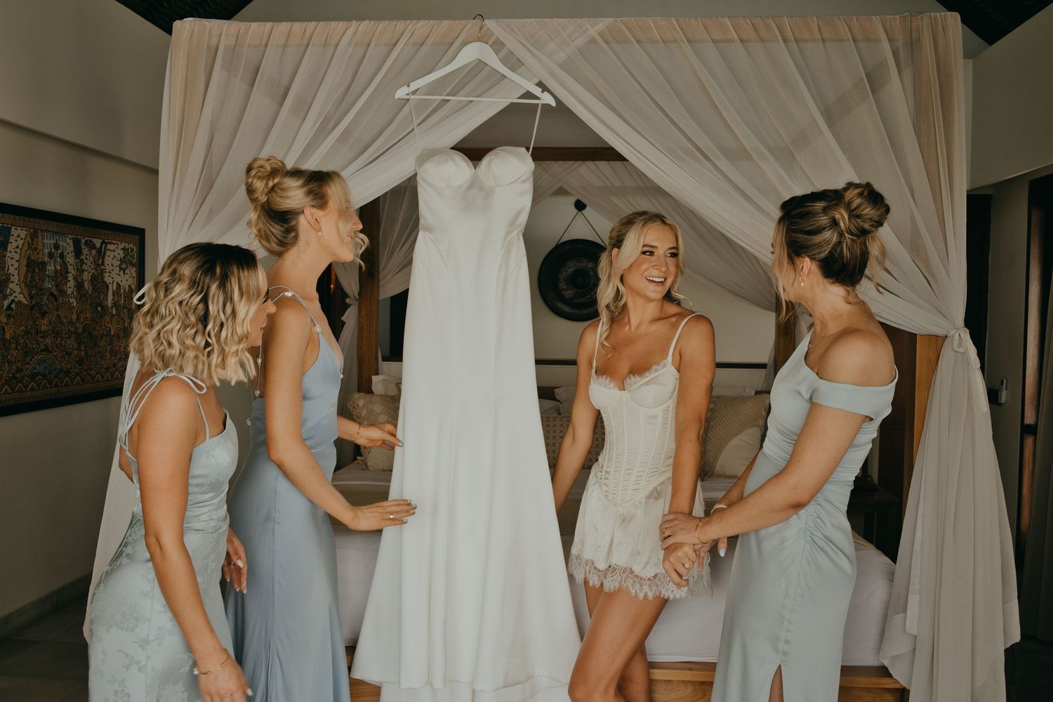 Bride in white lingerie surrounded by three bridesmaids in blue dresses admiring hanging wedding gown.