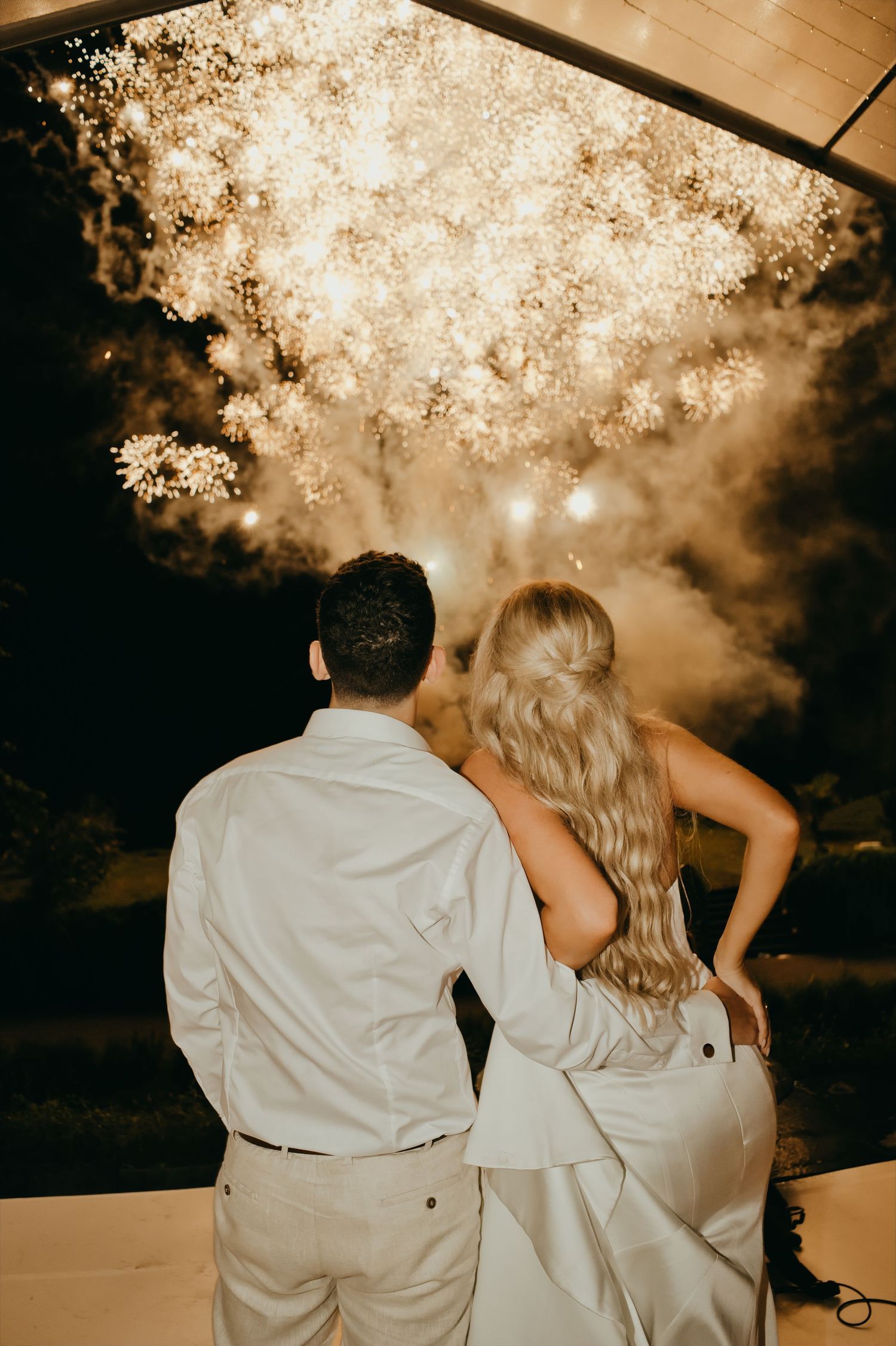 Newlyweds watch stunning fireworks display together at outdoor evening wedding reception.