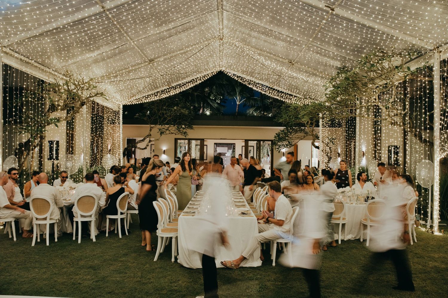 Elegant wedding reception under fairy light canopy with long white tables and dancing guests.