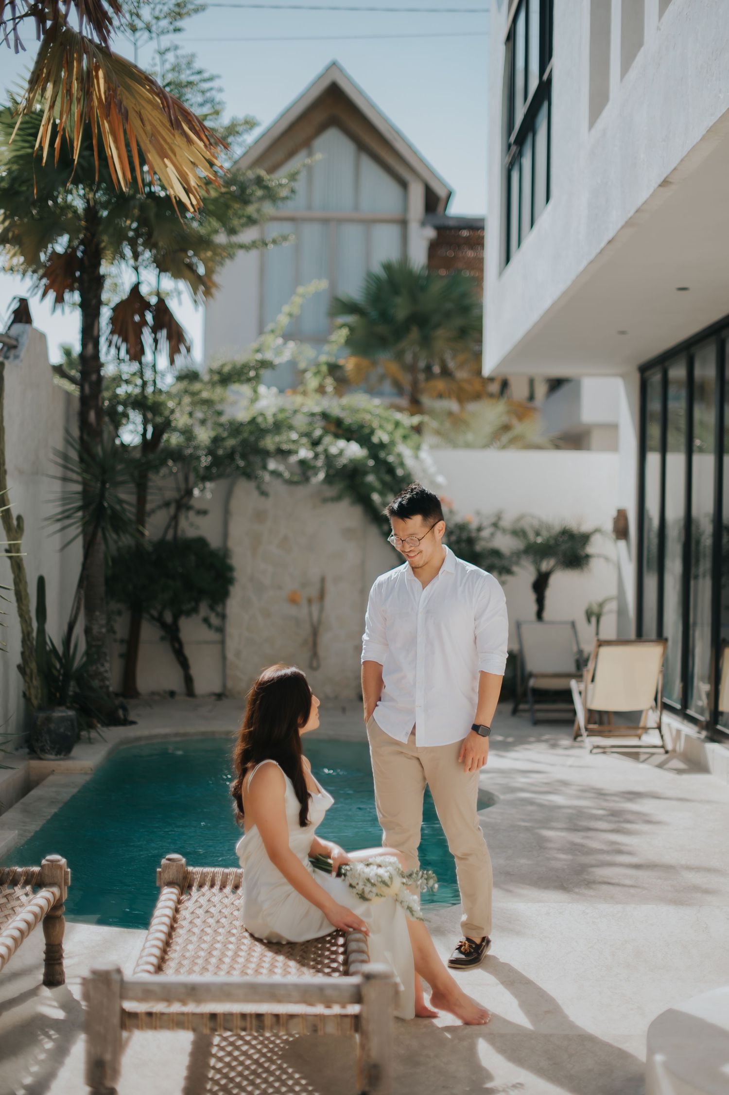 Couple enjoying a sunny poolside moment at a modern villa, woman seated on lounge chair holding flowers, man standing nearby.