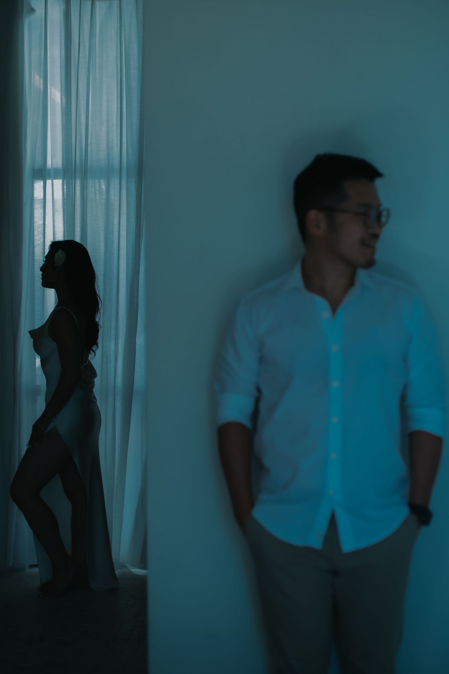 A man in a white shirt leans against a wall while a woman's silhouette stands by sheer curtains in dim blue light.
