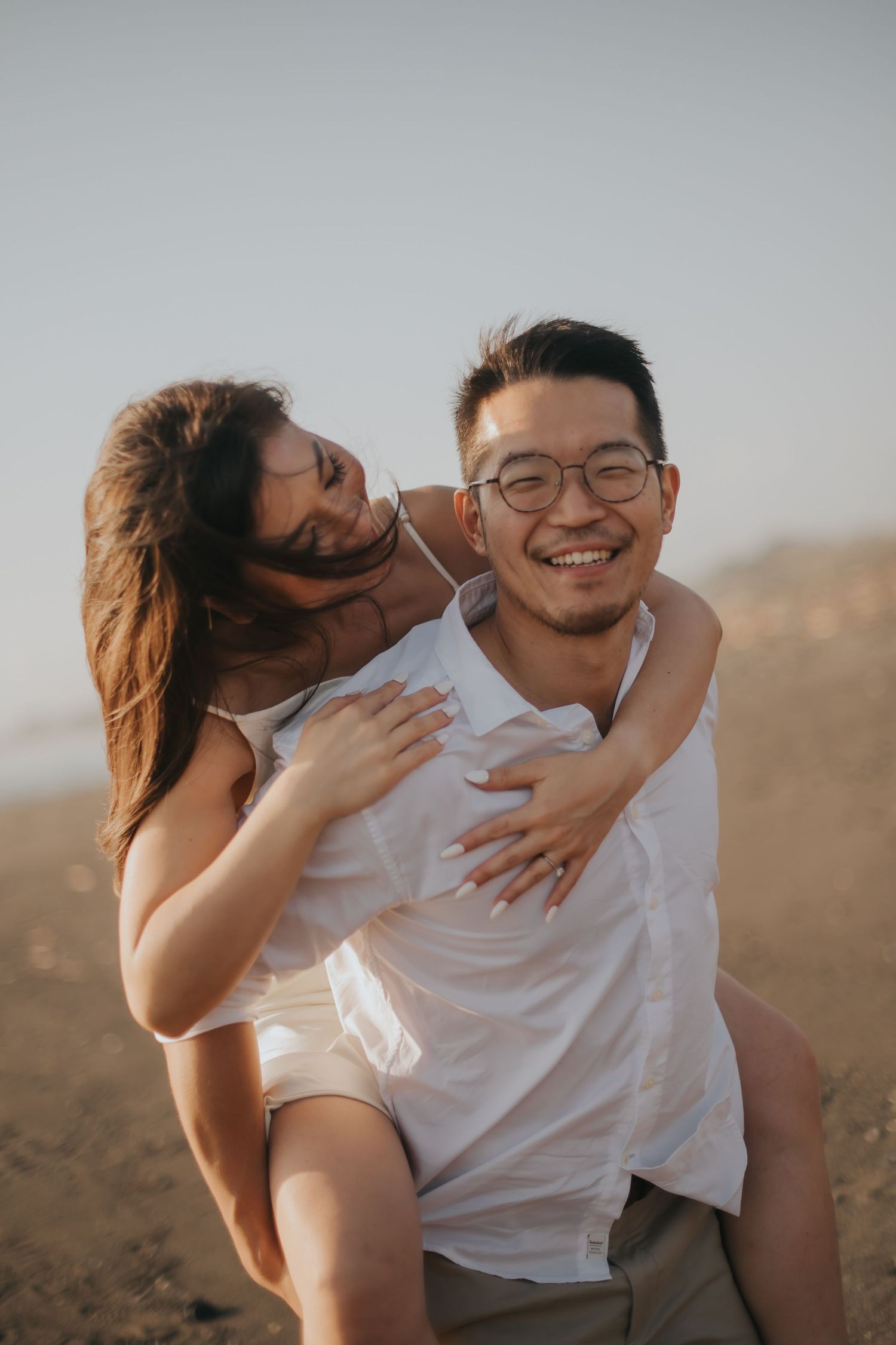 A smiling man gives a laughing woman a piggyback ride on a sandy beach at sunset.