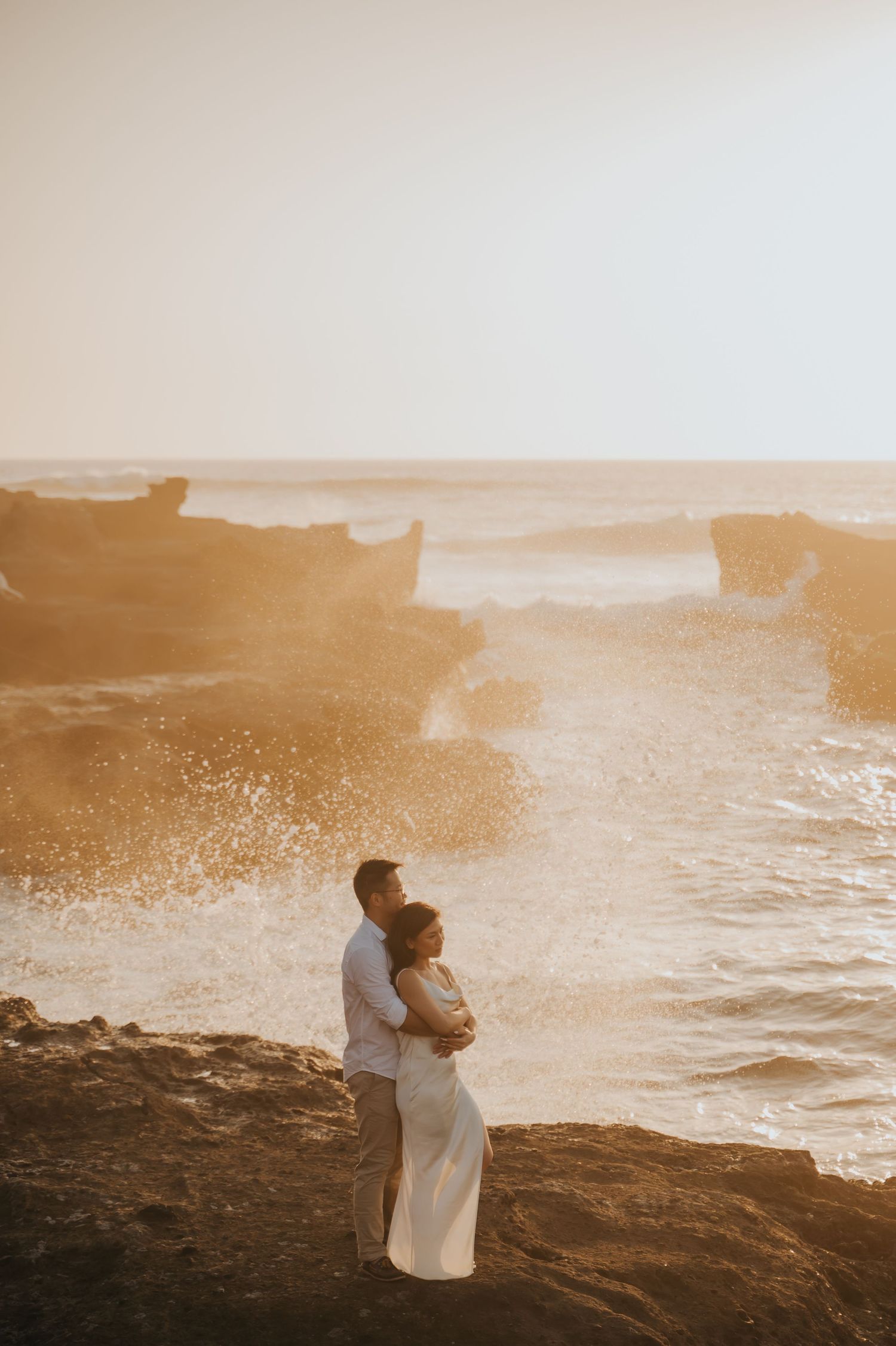 Couple embracing on rocky coastline at golden sunset with dramatic ocean waves crashing behind them.