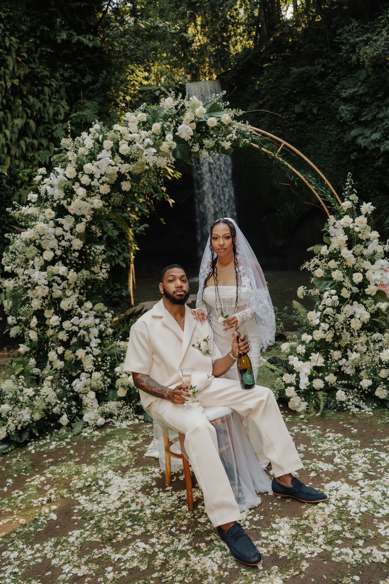 Newlyweds celebrating with champagne under white floral arch at tropical Bali waterfall wedding.