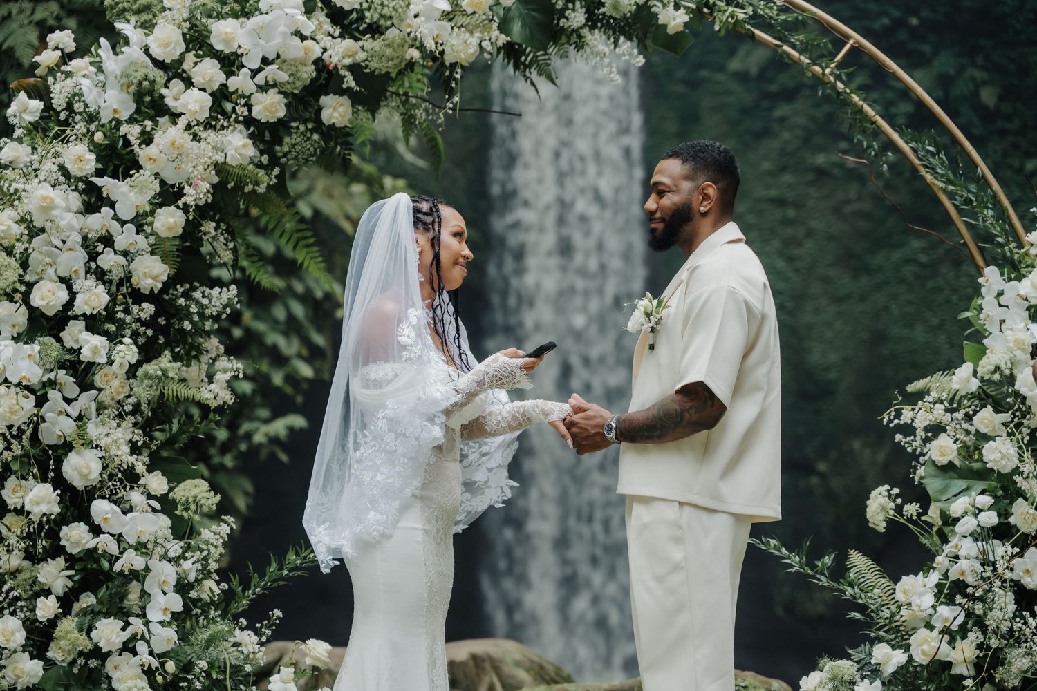 Couple exchanging vows under white floral arch with waterfall background at Bali wedding.