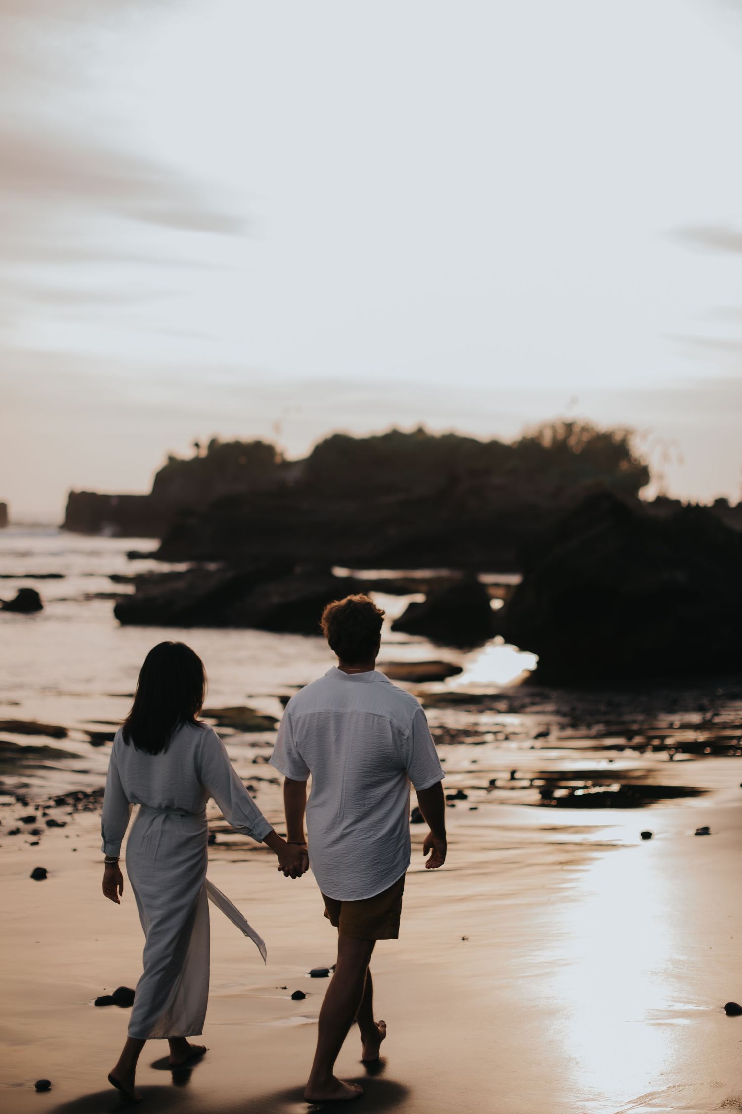 Two people walk along a beach at sunset with dark silhouettes against a golden sky and ocean backdrop.