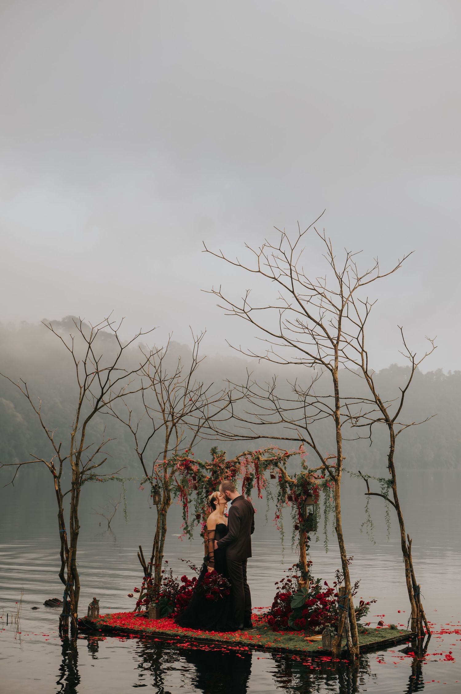 A misty lake ceremony setup with bare trees and floating red petals on water.