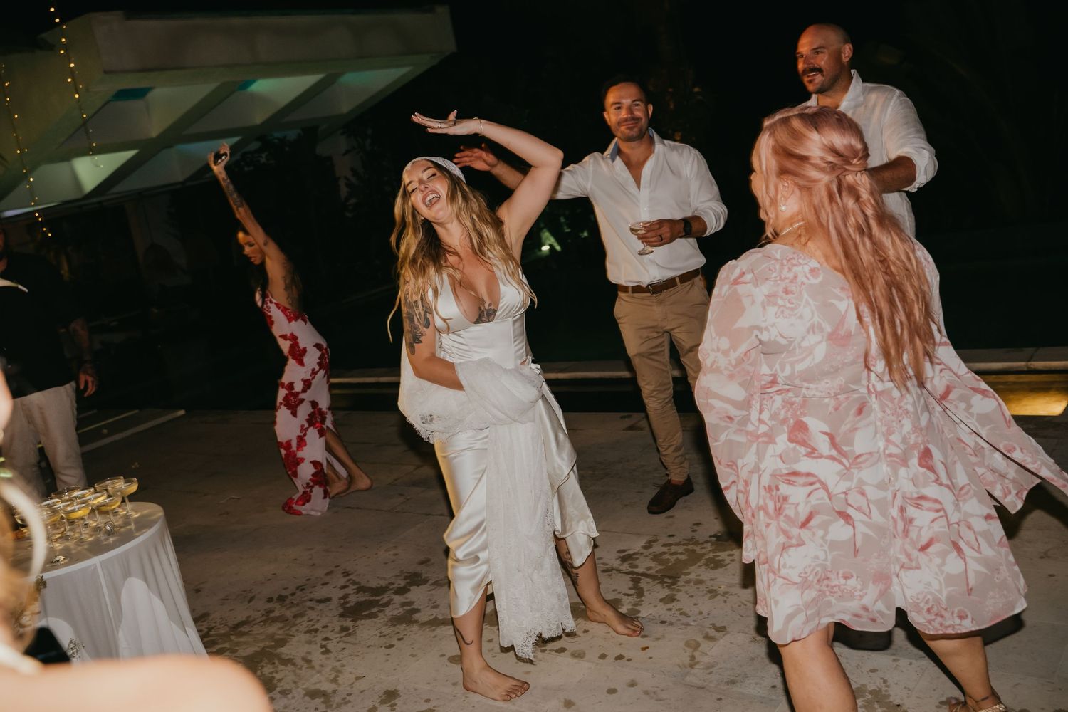 Barefoot bride dances barefoot with guests at a lively outdoor evening wedding reception.