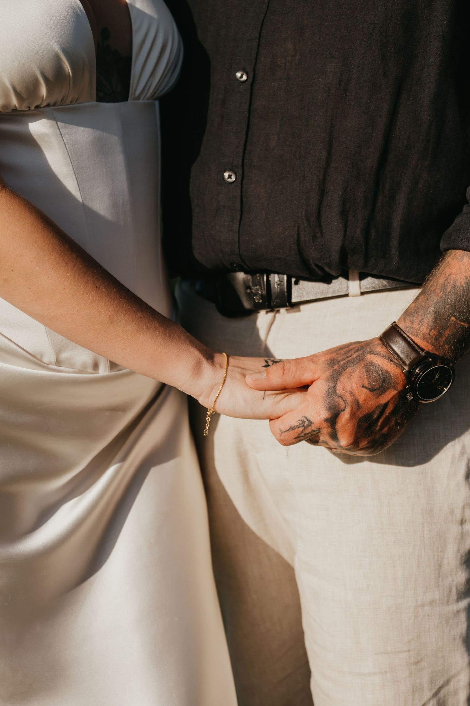 Close-up of bride and tattooed groom holding hands, groom wearing leather watch and black linen shirt.