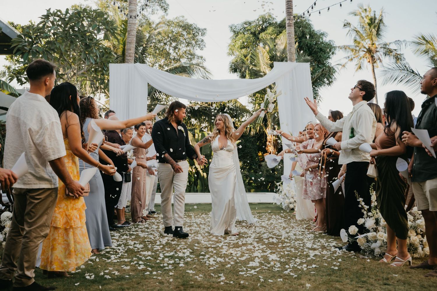 Newlyweds walk down aisle as guests toss white confetti under tropical outdoor wedding arch at sunset.