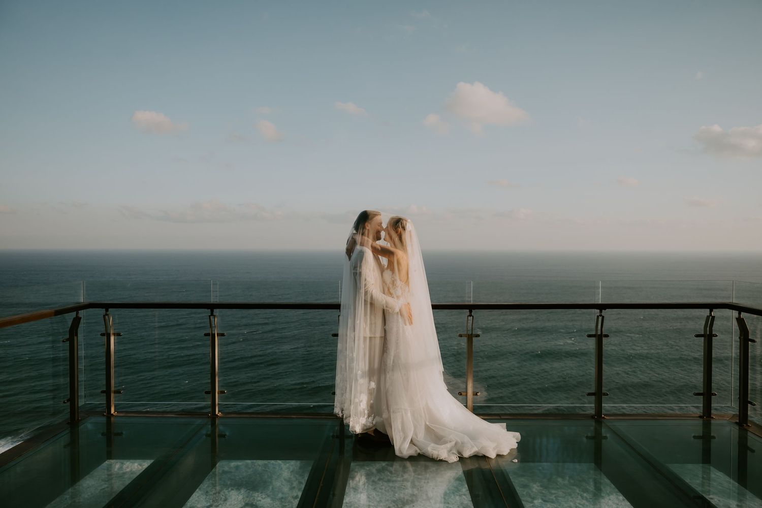 Two brides in white gowns share a kiss on a glass balcony overlooking the ocean on their wedding day.