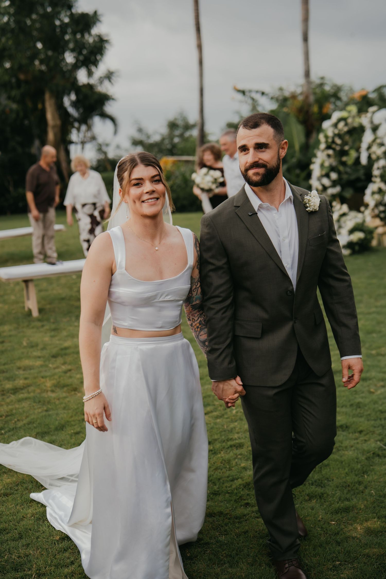 Newlywed couple walking hand in hand down garden aisle smiling after outdoor wedding ceremony.