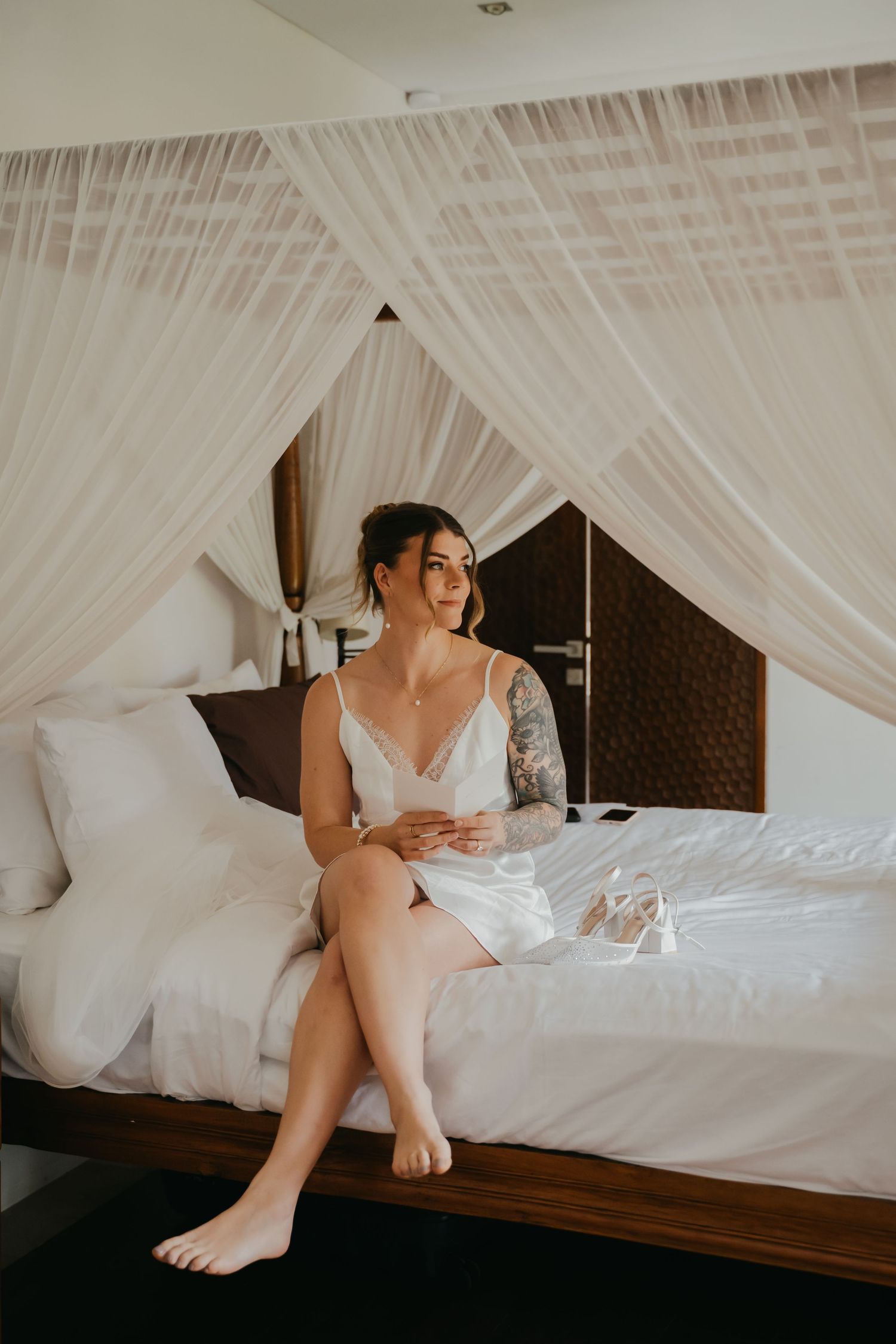 Bride in white silk slip dress sits barefoot on canopy bed gazing thoughtfully, tattoos visible on arm.