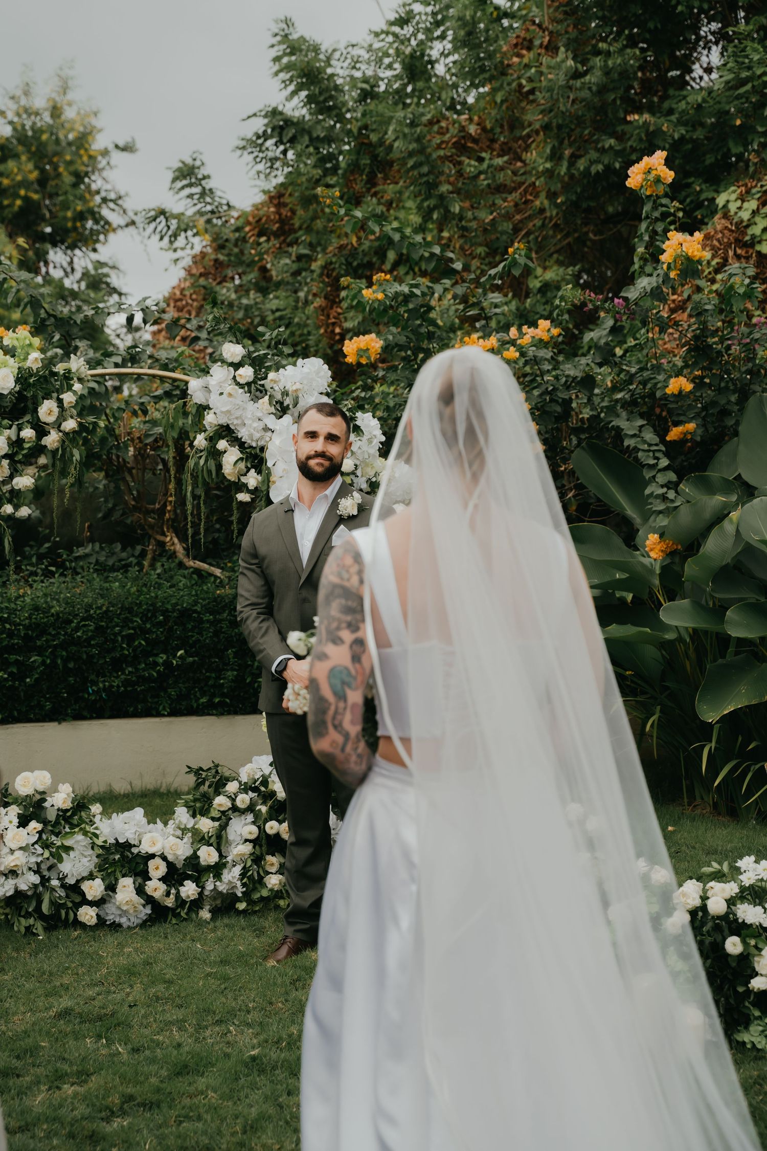Groom gazing at veiled bride before floral arch with white and yellow blooms.